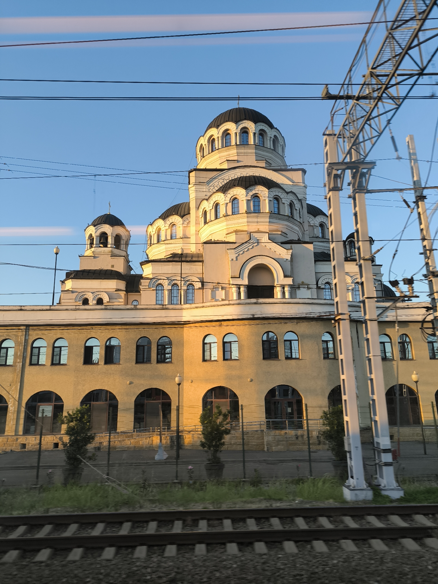 A grand, white-and-gold Orthodox church with multiple domes dominates the frame, bathed in the warm glow of what appears to be late afternoon or early evening sunlight. The building's architecture is ornate, featuring arched windows and decorative detailing. In the foreground, train tracks run horizontally across the bottom of the image, with overhead power lines and support structures creating a latticework pattern against the clear blue sky, which shows faint streaks of pink and orange from the setting sun.
To the left of the church, a smaller bell tower with a dome and a cross is visible. The lower section of the church building is a pale yellow, also adorned with arched windows. A black metal fence separates the building from the train tracks, with sparse green grass and a few small trees growing behind it. The sunlight creates strong shadows and highlights on the church, emphasizing its architectural features.
The scene is static, suggesting a view captured from a moving vehicle, likely a train, given the presence of the tracks and overhead lines. The overall mood is serene and majestic, with the golden light casting a sense of peace over the imposing structure. No people are visible in the image. No text is discernible. The location context of Sochi, Russia, places this scene in a coastal city known for its distinct architecture and its role as a major transportation hub.