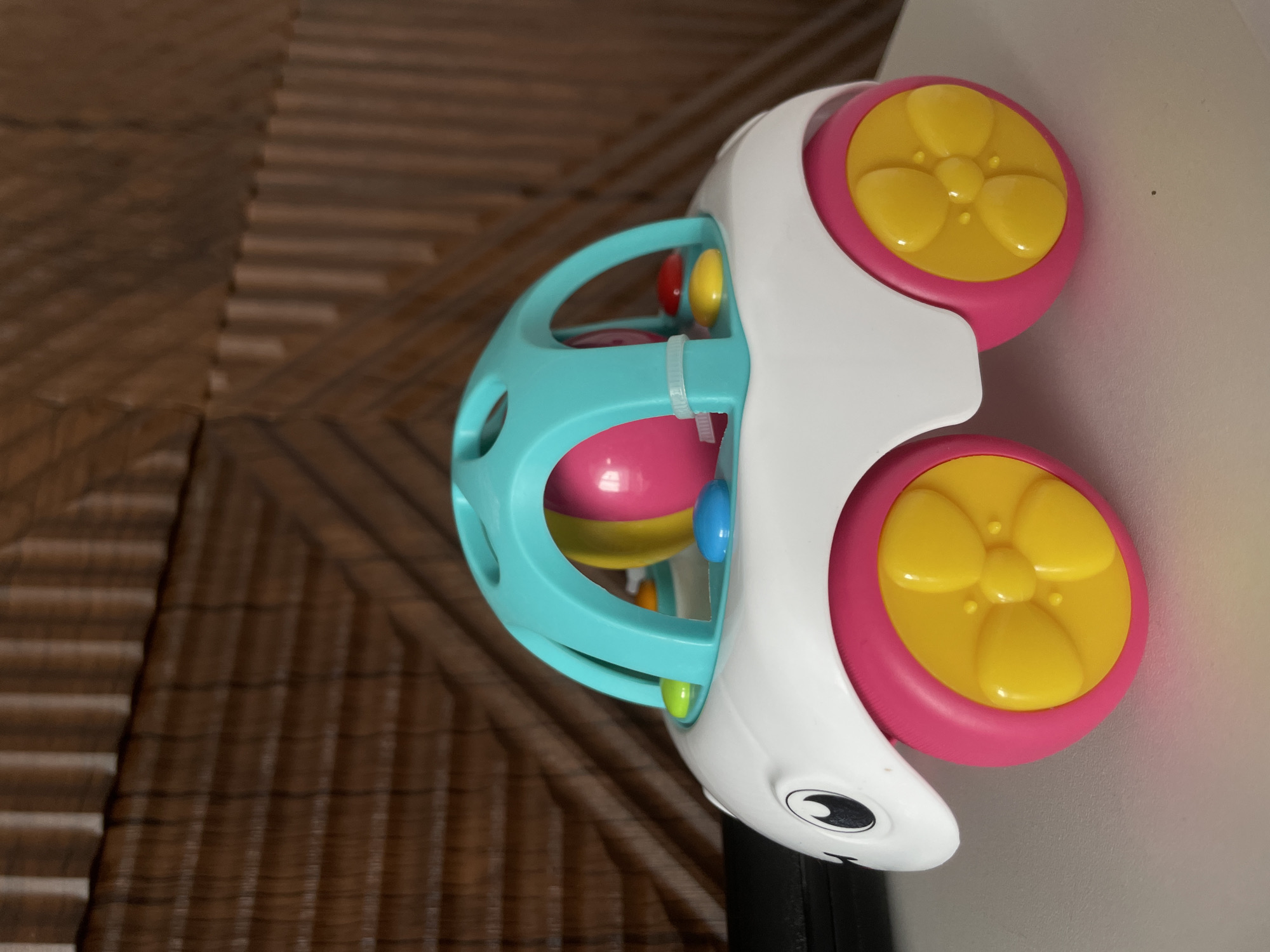 The image shows a colorful baby toy with a white, animal-shaped body and two pink wheels with yellow centers. A teal cage-like structure surrounds a ball filled with various colored balls. The toy is resting on a brown, woven surface and part of a white wall. It is not possible to identify any specific details relating to Средний Постол, Russia from the image.