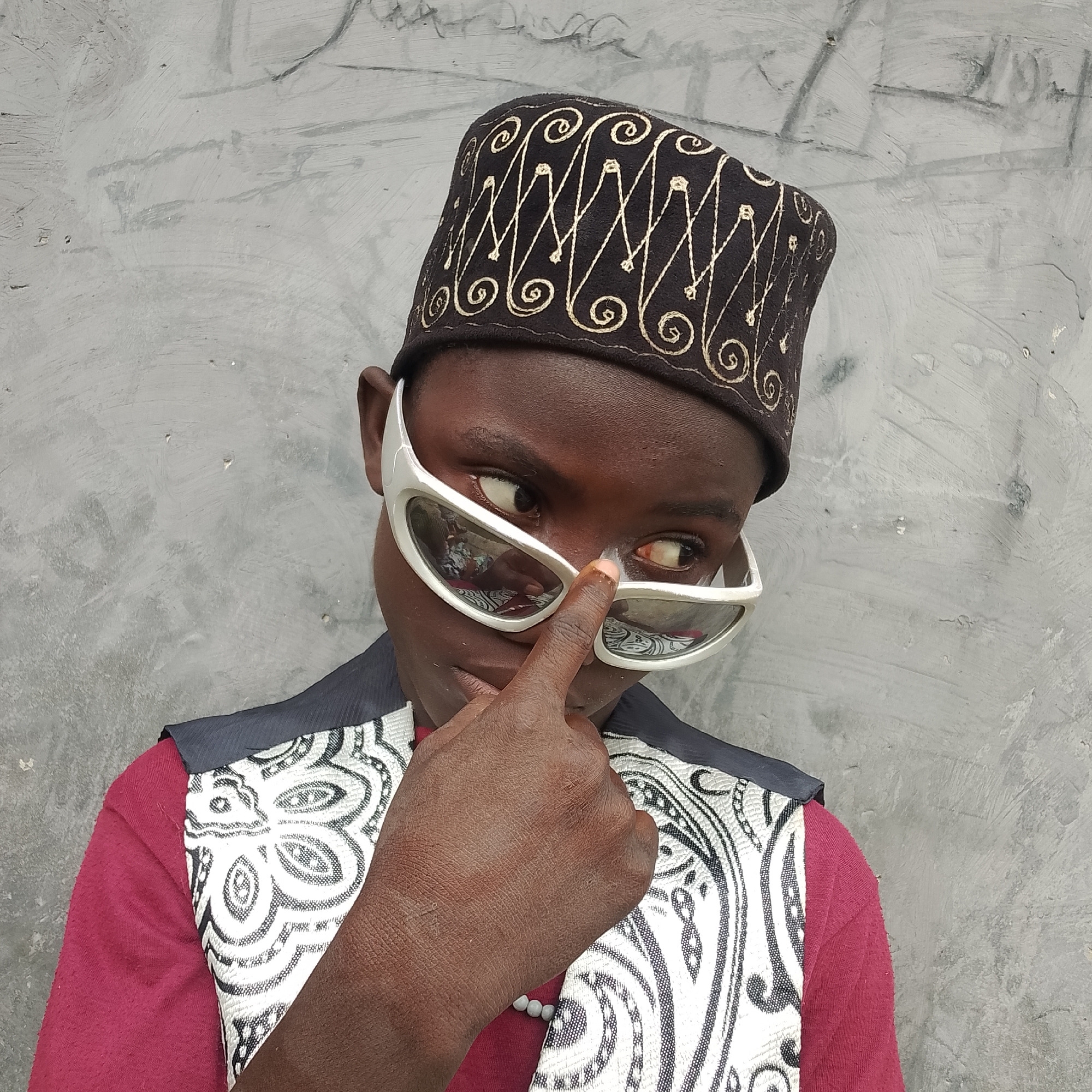 A close-up, waist-up shot captures a young Black individual wearing a maroon t-shirt, a grey vest with a white, black, and grey patterned fabric overlay, and a dark cap with gold embroidered swirls and diamond shapes. The individual is wearing large, white, angular sunglasses and is looking off to the right of the frame, with their right index finger touching the bridge of their nose, slightly pushing up the sunglasses. The background is a textured grey wall with some visible scuffs and marks, suggesting an outdoor urban setting, possibly Ado, Nigeria. The lighting is diffused, casting soft shadows and highlighting the textures of the clothing and the wall.