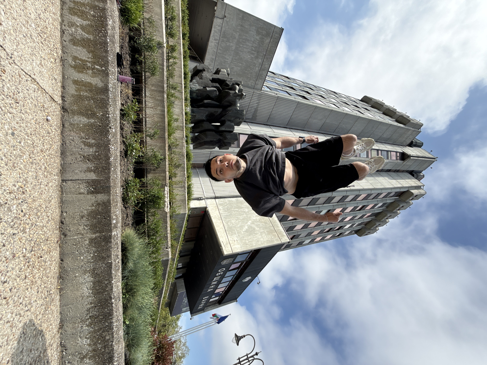 A medium-shot captures a man appearing to float in front of the Varna Municipality building in Varna, Bulgaria. The man, wearing a black t-shirt, black shorts, and light-colored sneakers, is positioned horizontally, seemingly suspended in mid-air with the building as a backdrop. The modern building has a stepped, rectangular design. A black sculpture stands to the side of the building. The sky is visible with wispy clouds behind the building. A concrete ledge with some vegetation is in the lower left of the frame. The image appears to be rotated counterclockwise. Flags fly in front of the building.