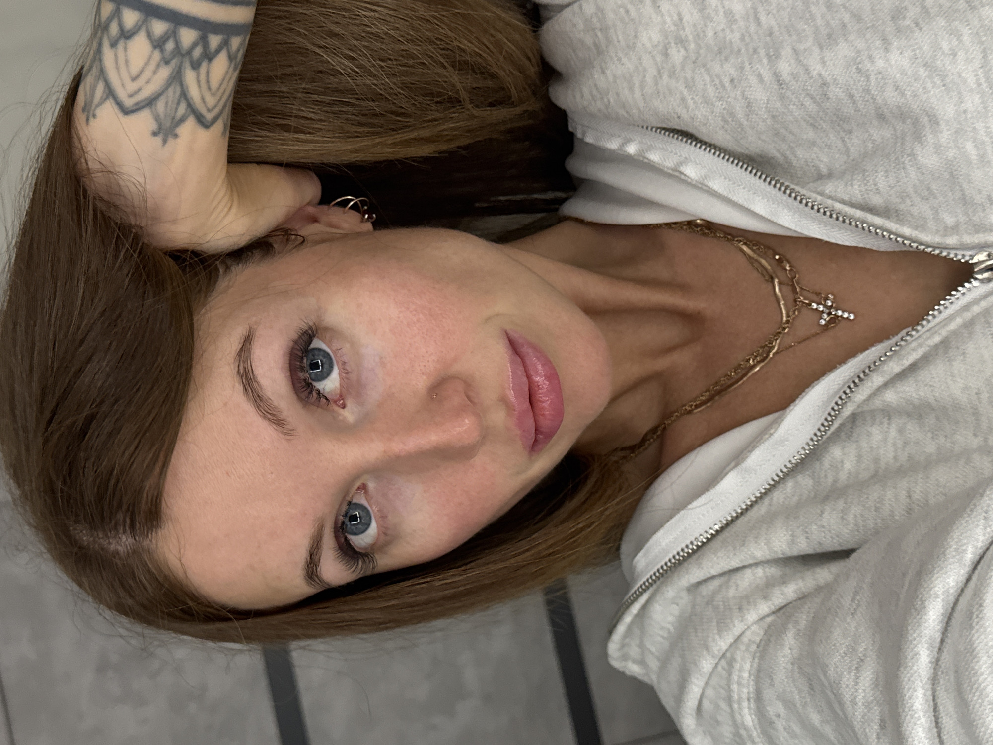 A fair-skinned woman with light brown hair and blue eyes is depicted in a selfie. She is wearing a light gray zip-up hoodie, partially unzipped to reveal a gold necklace with a cross pendant. She has a tattoo on her left arm. A small gold earring is visible in her left ear. The background appears to be a gray tiled surface. The image was taken indoors, and no information suggests the location is in Chaikovsky, Russia.