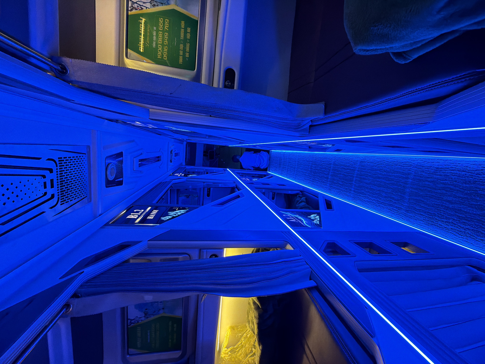The image captures the interior of what appears to be a modern sleeper bus or capsule accommodation, likely in Vietnam, given the visible text. The dominant feature is the striking blue LED strip lighting that lines the ceiling and floors of the narrow aisle, creating a futuristic and ambient atmosphere.
The scene shows a row of individual sleeping pods or beds, each equipped with curtains for privacy. One pod on the left side is labeled "BL2 HANOI." In the background, a person is visible sitting upright inside one of the pods, seemingly resting or engaged with a device, illuminated by the blue light. Another pod on the right shows a warm, yellow interior light, contrasting with the blue, suggesting a different lighting option within the individual compartments.
A prominent green and white sign is visible at the top, displaying Vietnamese text: "ĐÌNH HOÀI Limousine," indicating a premium bus service. Below this, "TỔNG ĐÀI ĐẶT VÉ" (Booking Hotline) is printed, followed by two phone numbers: "1900 888 666" and "0916 949 789." Further down, tour routes are listed: "TOUR HÀ NỘI - THANH HOA" (Tour Hanoi - Thanh Hoa) and "DA NANG = HOI AN" (Da Nang = Hoi An). The mention of Đà Nẵng and Hoi An aligns with the provided location context.
The design of the pods is sleek and enclosed, featuring textured surfaces and possibly ventilation grilles. The general absence of natural light suggests it is either nighttime or the bus is designed to block outside light for passenger comfort during travel. The image highlights a modern travel experience, emphasizing comfort and privacy on long journeys.