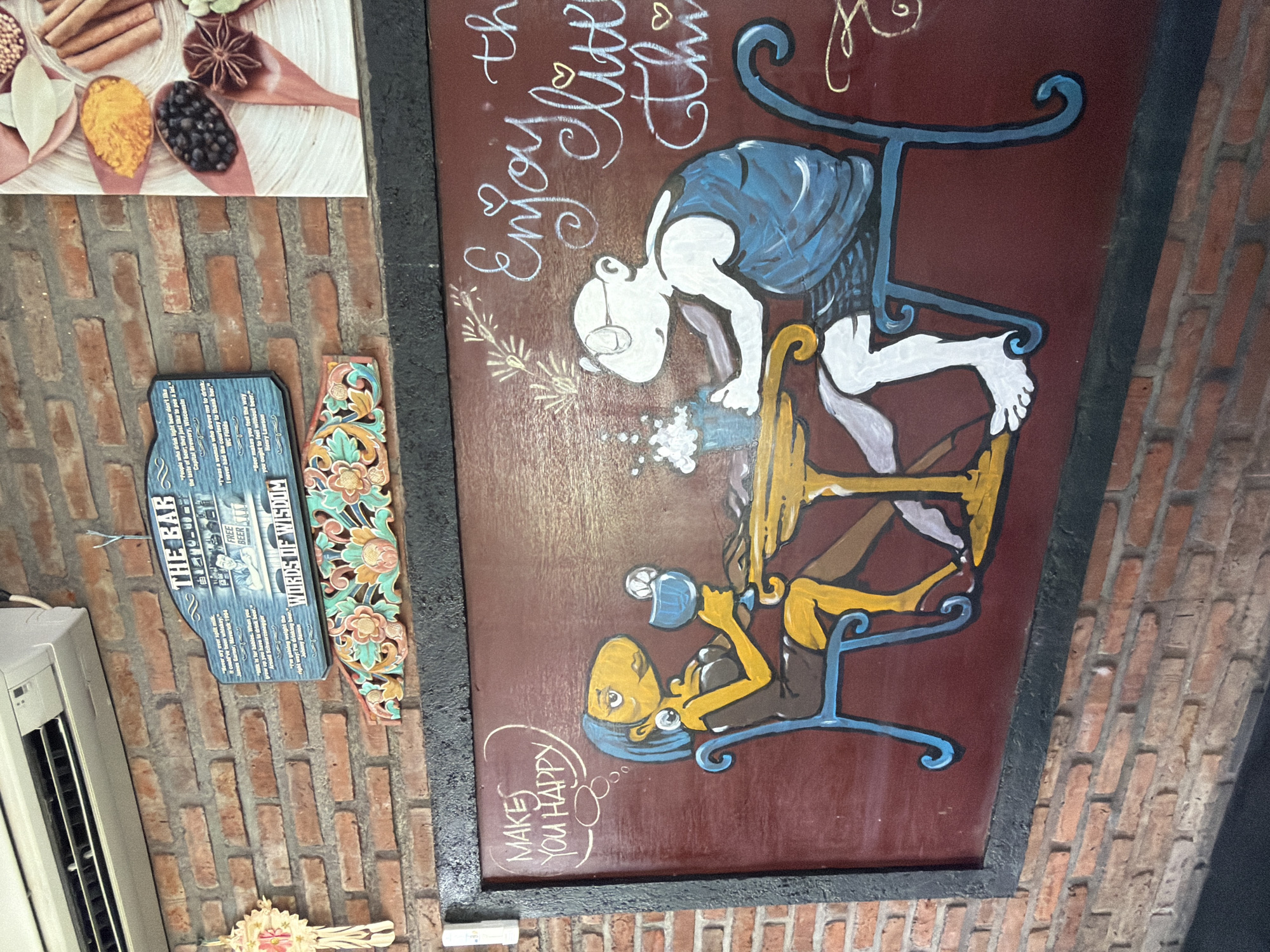 The image shows a mural on a brick wall in North Kuta, Indonesia. The mural depicts two stylized figures sitting at a table, with text above them. The figure on the left is female with blue hair, yellow skin, and large earrings, holding a blue cocktail. The figure on the right is male with pale skin, glasses, and a blue garment. Above the woman, text in a thought bubble reads "MAKES YOU HAPPY". To the right of the male figure, in cursive white lettering, is the phrase "Enjoy the little thi". The background of the mural is a dark red. Above the mural, a blue sign with white lettering hangs on the brick wall and reads "THE BAR WORDS OF WISDOM". To the left of this sign is a white object, possibly an air conditioner, and a decorative wooden carving with floral patterns hangs below it. The overall setting appears to be an indoor establishment with exposed brick walls.