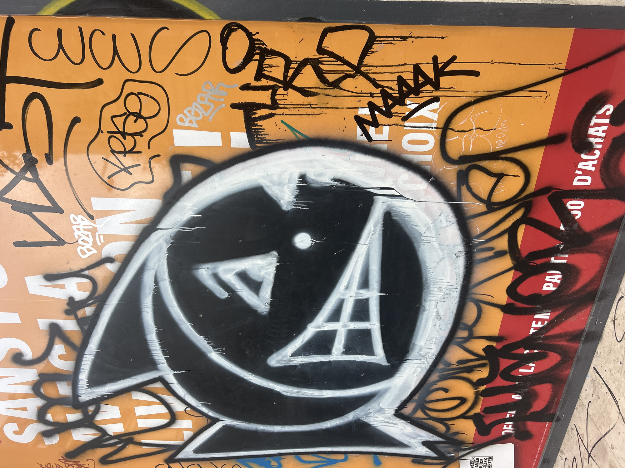 The image depicts graffiti on an orange and red surface. The graffiti includes writing in black, as well as a black and white design in the center resembling a stylized face with a triangle and a ladder-like shape within it. Other markings and tags are present on the surface, some obscuring the original background, which appears to be a poster or advertisement with text written in French. The environment appears to be an urban area.