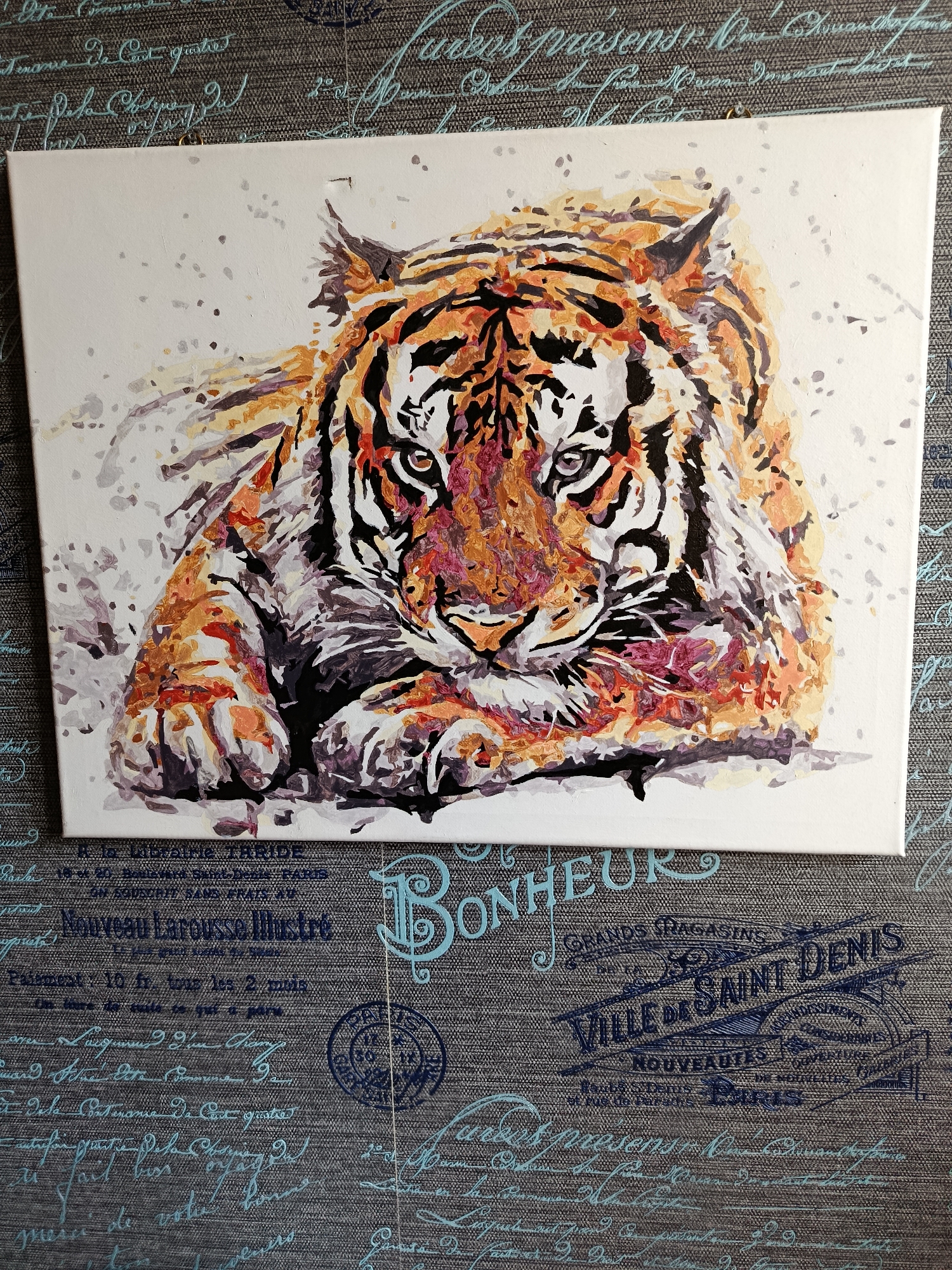 The image displays a paint-by-numbers canvas depicting a tiger's face. The canvas is placed on a textured dark gray fabric with a pattern of light blue script and circular stamps. The tiger's fur is rendered in shades of orange, black, white, and purple. Its eyes are partially closed, and its gaze is directed forward. The painting is presented in a top-down view. The fabric beneath the canvas appears to be a tablecloth or a similar textile. The setting is indoors, as indicated by the lighting and the indoor environment suggested by the fabric. There are no people or animals present other than the painted tiger. The city of Apatity, Russia, is not depicted or implied in this image.