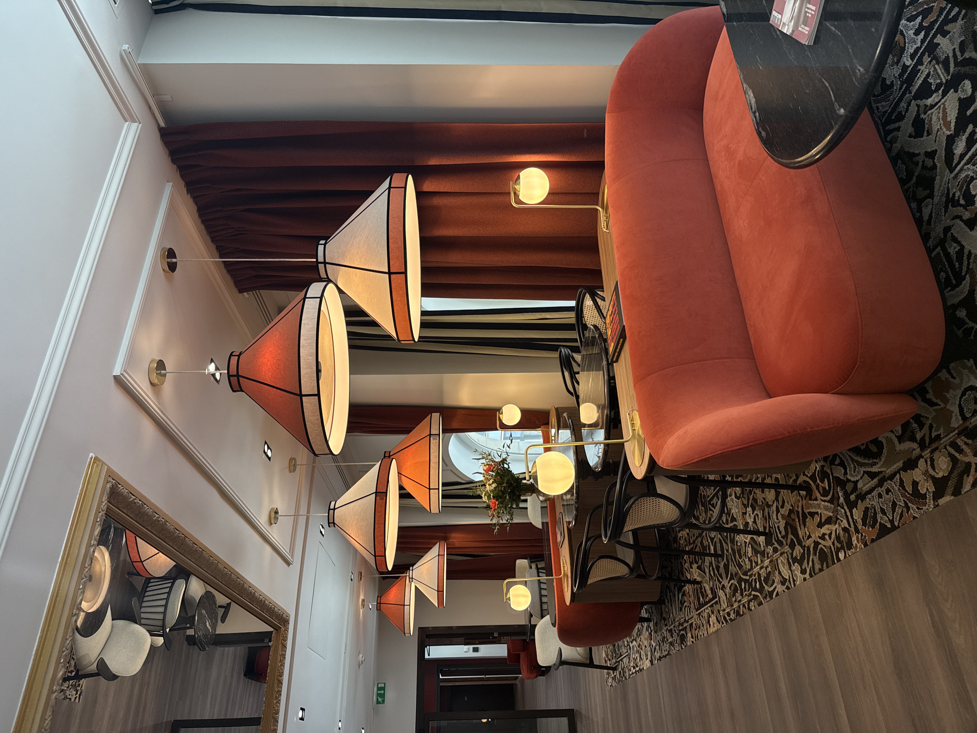 The image shows a section of a restaurant interior. A large, curved orange sofa is positioned along the right side of the frame, next to a small black table. The floor is a light wood finish, with a decorative patterned rug underneath the seating area. A row of hanging light fixtures runs down the left side of the image, each with a distinctive conical shade that has an orange tint. The lights are evenly spaced along the wall, which is white and features ornate molding. Along the top of the image, rust-colored curtains provide a warm accent. In the distance, a doorway leads to another section of the restaurant, where additional seating is visible. The scene appears calm and unoccupied, capturing a moment of stillness within the restaurant.