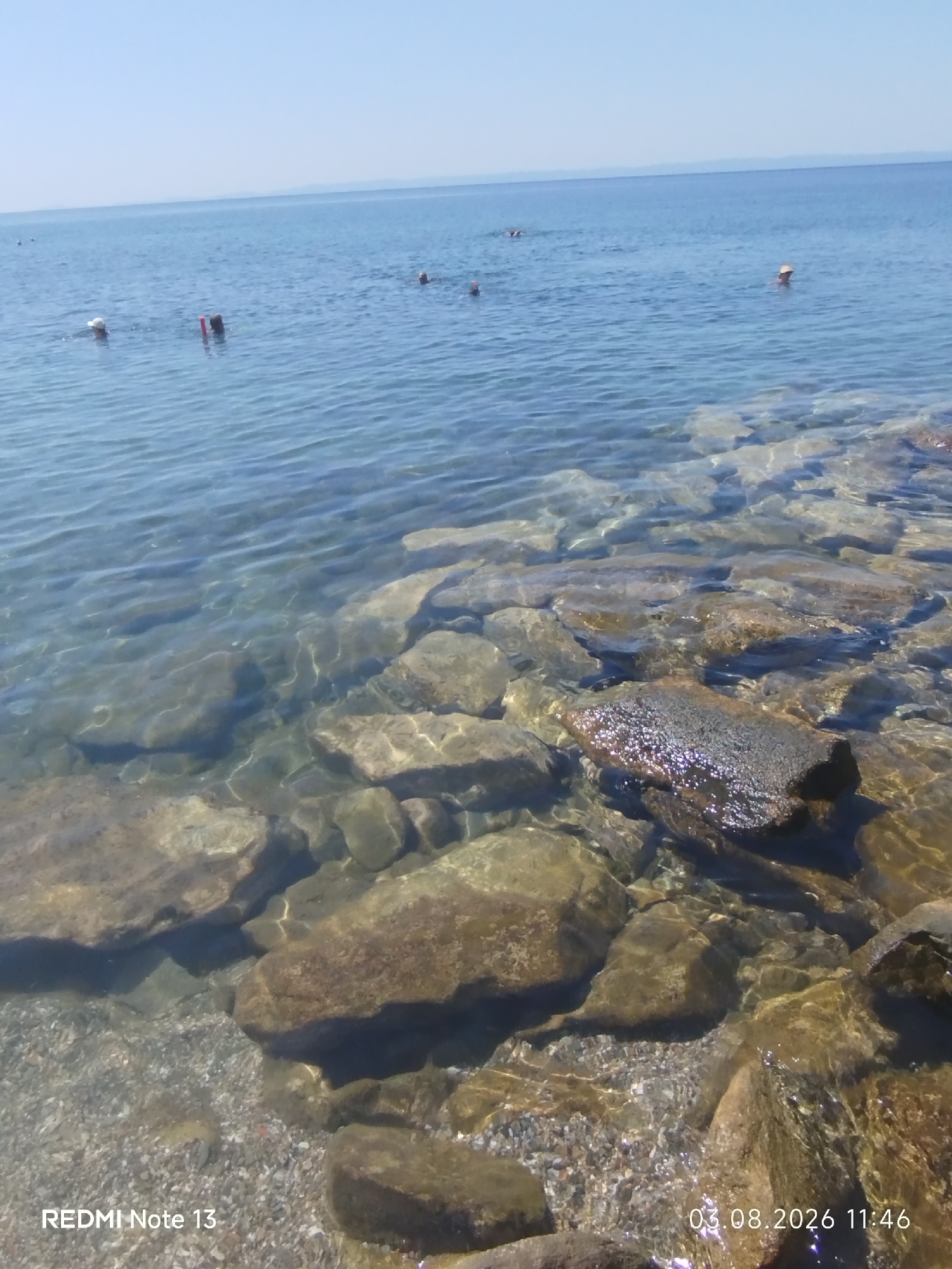 The image depicts a coastal scene with clear, shallow water revealing a rocky seabed. In the distance, several people are visible in the sea, some appearing to be swimming or wading. The water is calm, with gentle ripples on the surface reflecting the bright sky. The shoreline is composed of numerous stones and pebbles. The overall setting suggests a natural beach or cove. The timestamp on the image indicates it was taken on August 3, 2026, at 11:46.