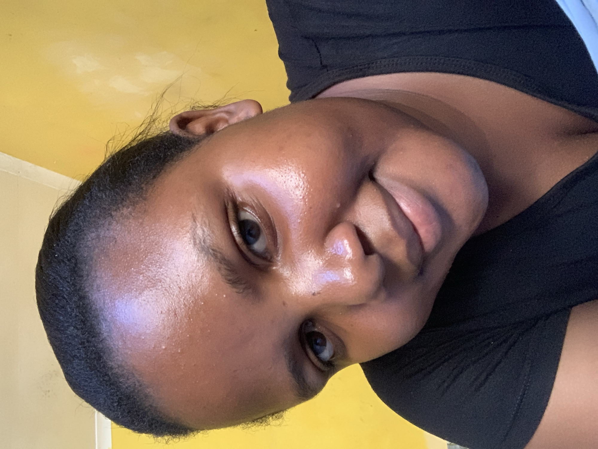 A close-up headshot of a dark-skinned woman with black hair pulled back, positioned against a yellow wall. She is wearing a black top. The location is unspecified and it is unknown if it is in Dar es-Salaam, Tanzania.