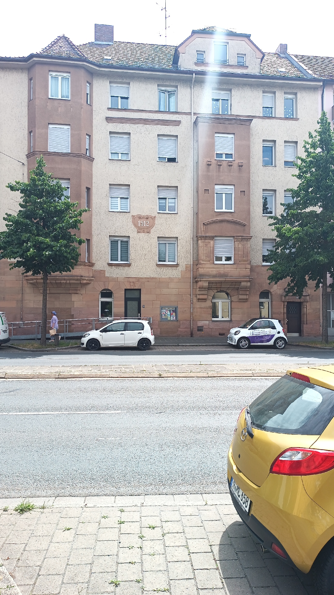 The image shows a street view in Nuremberg, Germany, featuring a building with a stone facade and an inscription reading "1912". A white car and a purple and white smart car are parked on the street in front of the building. A person is walking on the sidewalk next to a tree. The street is paved with asphalt, and the sidewalk is made of cobblestones. The sky is overcast.