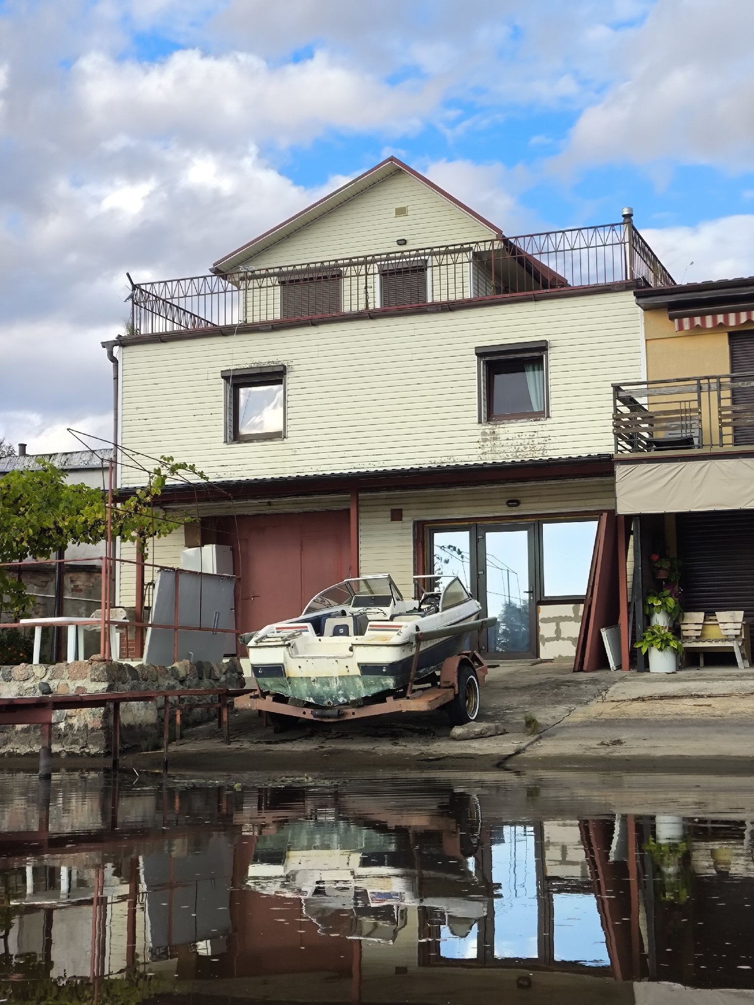 The image depicts a waterfront scene in Ushakovo, Russia. A light-colored, two-story building with horizontal siding dominates the frame. The building features a balcony with a dark metal railing on the upper level and a large red double door on the ground floor. To the right of the main building, a section of a yellow building with a striped awning is visible. In front of the building, a boat on a trailer rests on the shore. A wooden pier extends into the water from the left side of the image. The water is calm and reflects the building and the sky, which is partly cloudy with patches of blue. Several potted plants and wooden benches are situated near the building. The overall impression is one of a potentially residential or commercial property located by a body of water.