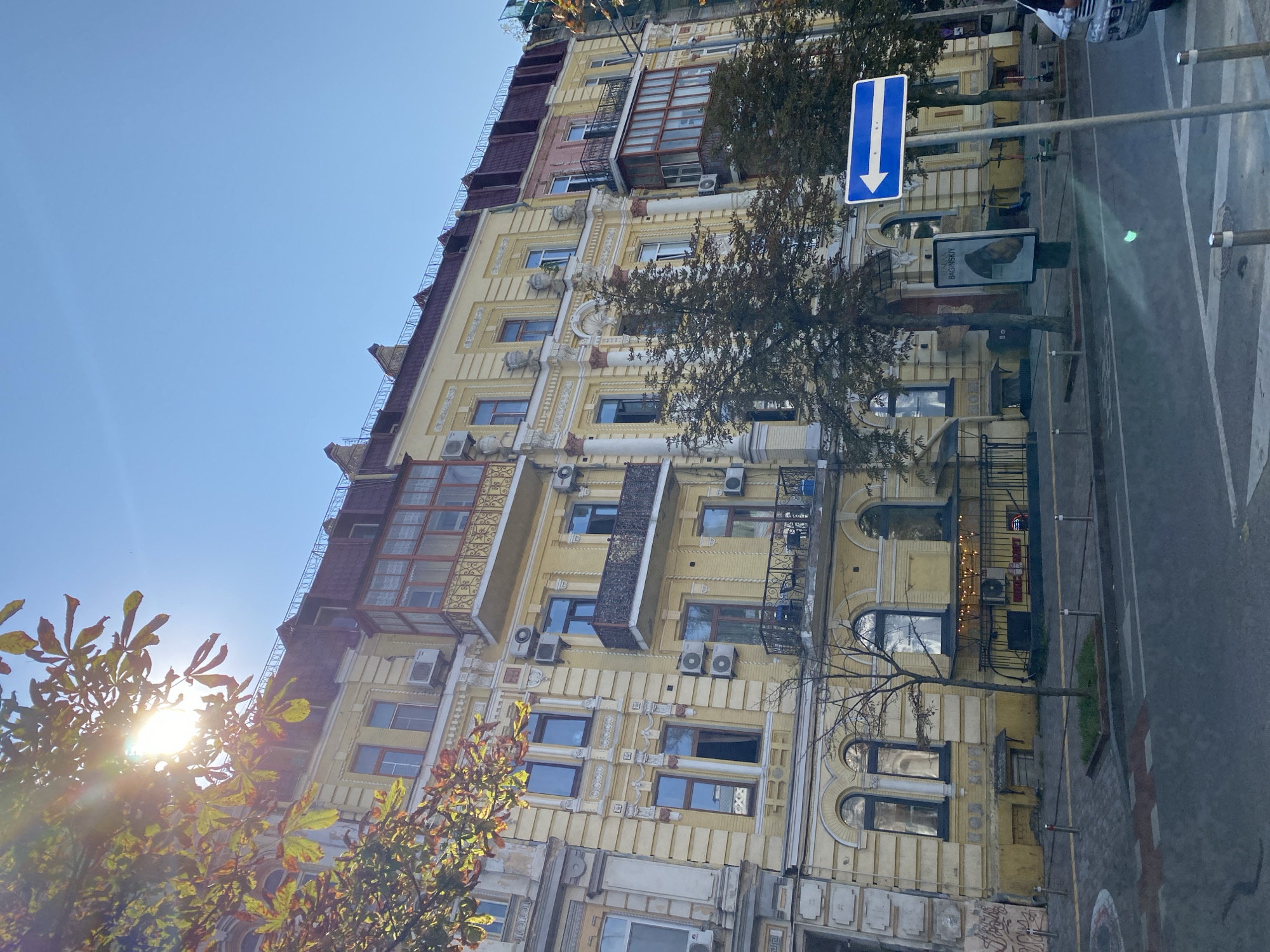 The image shows a yellow apartment building on a sunny day. The building has multiple stories and features decorative architectural details, balconies, and numerous windows. Trees with autumn foliage are visible in the foreground and near the building. A blue and white directional sign indicating a left turn is posted on the right side of the image, next to the building. The street in front of the building has parking meters, a sidewalk, and a road with visible lane markings. There is some graffiti on the lower left portion of the building. The sky is clear and blue.