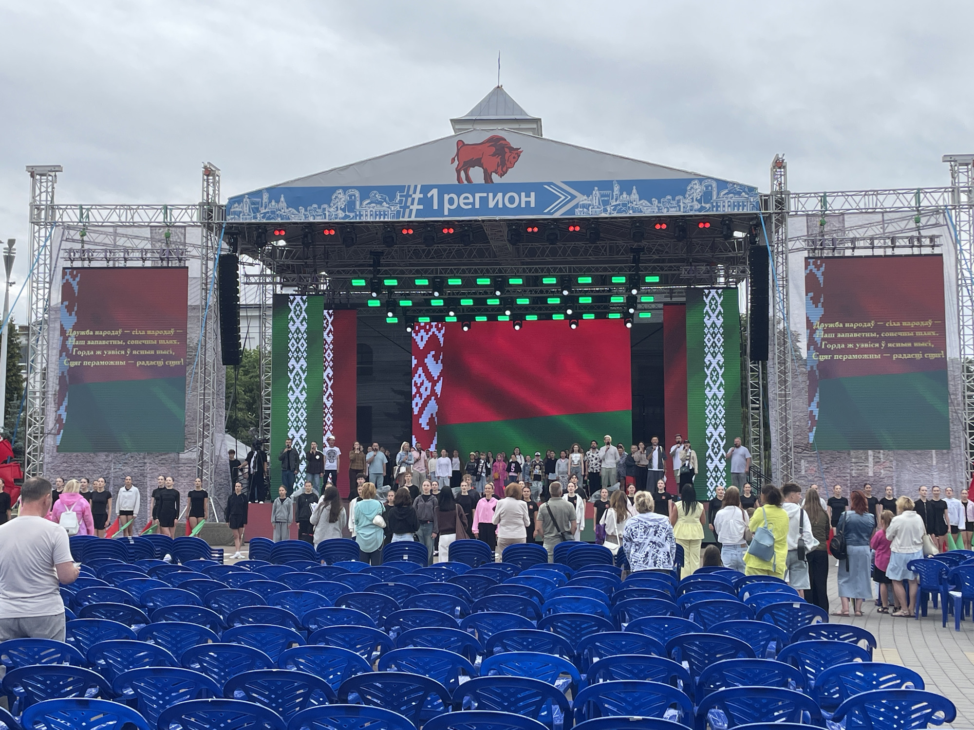 A large outdoor stage is set up in an urban area, likely for a public event. The stage backdrop features a prominent red bull logo atop a banner that reads "#1 Регион" ( "#1 Region" in Russian), suggesting a regional celebration or competition. The stage itself is adorned with large LED screens displaying the flag of Belarus, which is a red and green pattern with a white ornamental pattern on the left side. The screen is flanked by vertical banners also featuring the Belarusian ornamental pattern.
A crowd of people, a mix of adults and children, are gathered in front of the stage, with many standing and some seated in rows of blue plastic chairs in the foreground. Some individuals appear to be performers or organizers, dressed in various attire ranging from casual to semi-formal. There are speakers and lighting equipment visible on and around the stage, indicating a planned performance or presentation. The environment appears to be a public square or park, with a cloudy sky overhead. The overall scene suggests a festive or commemorative event taking place in Brest, Belarus, given the Belarusian flag and the "#1 Region" text, which could refer to a specific administrative region within Belarus.
