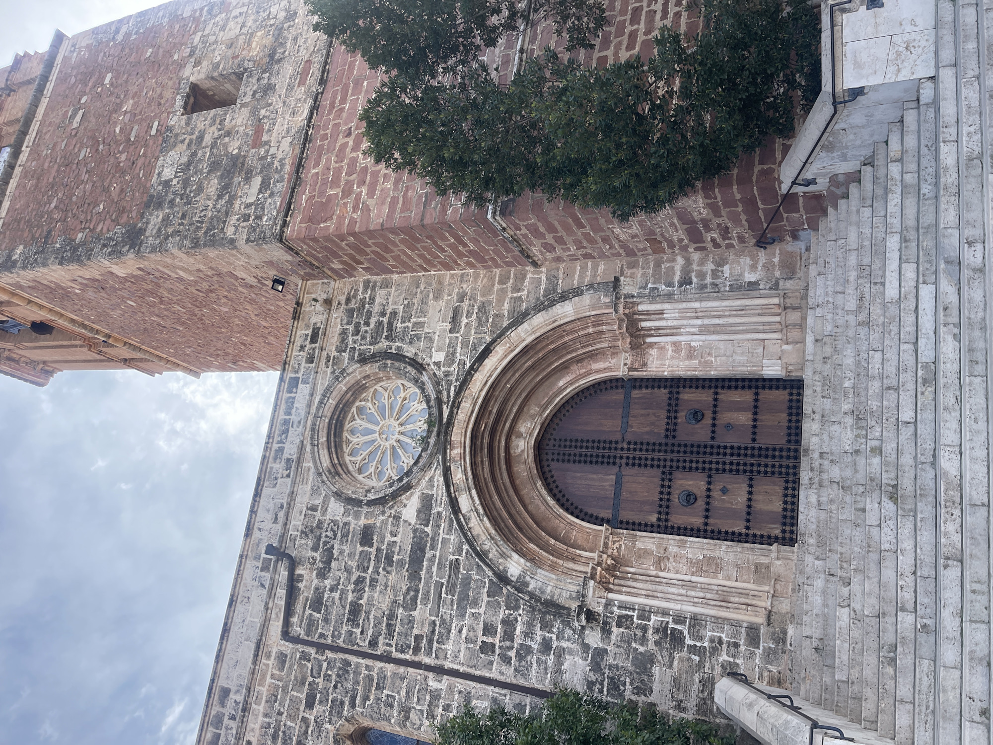 The image shows a low-angle view of the Monestir del Puig de Santa Maria, a historic monastery in El Puig de Santa Maria, Spain. The monastery is constructed with a mix of gray stone and red brick, showing clear variations in the building materials across different sections. A large arched wooden door is visible, ornamented with dark metal accents arranged in a grid pattern and circular knobs. To the left of the door is a circular window featuring a stained-glass rosette design. A set of stone stairs ascends the exterior, possibly leading to the entrance, while greenery partially obscures the upper sections of the building. The sky above is overcast, with a visible cloud cover.