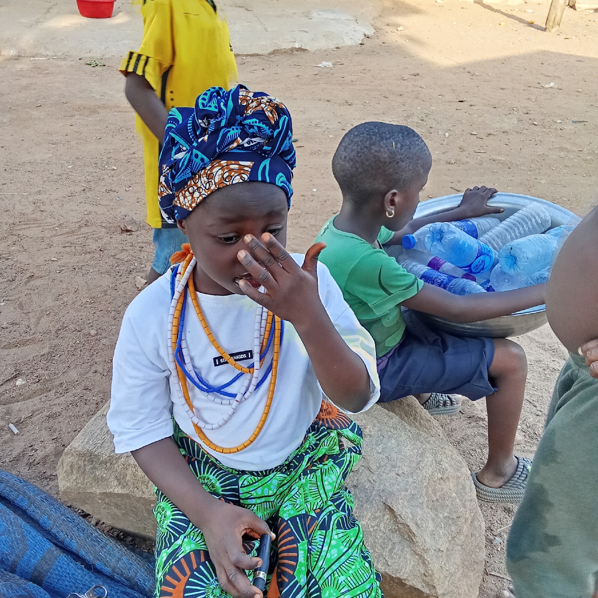 This image captures a daytime scene outdoors in Akwanga, Nigeria, featuring four children. In the foreground, a young child, likely a girl, is seated on a large light-colored rock. She wears a vibrant blue, orange, and white patterned headwrap, a long-sleeved white t-shirt, and a green skirt with a distinct black and orange pattern. Several colorful beaded necklaces in orange, white, and blue adorn her neck. Her right hand is raised to her face, partially obscuring her expression, while her left hand holds a small black object resembling a pen or cylindrical device.
Behind her, a second child, likely a boy, is also seated, wearing a green t-shirt and dark shorts, with a gold hoop earring visible in his left ear. He holds a large metal basin filled with numerous plastic water bottles, some clearly labeled "AQUA." Part of a third child in a green top is visible on the far right, and a fourth child in a yellow top and possibly blue trousers stands further back in the background.
The setting is characterized by a dry, sandy or dirt ground, suggesting an informal or rural outdoor environment. Shadows indicate bright sunlight. A red bucket is visible in the distant background. The visible text on the foreground child's shirt reads "I SEANKIDS |". The overall scene suggests daily life, possibly involving the sale or transport of water.