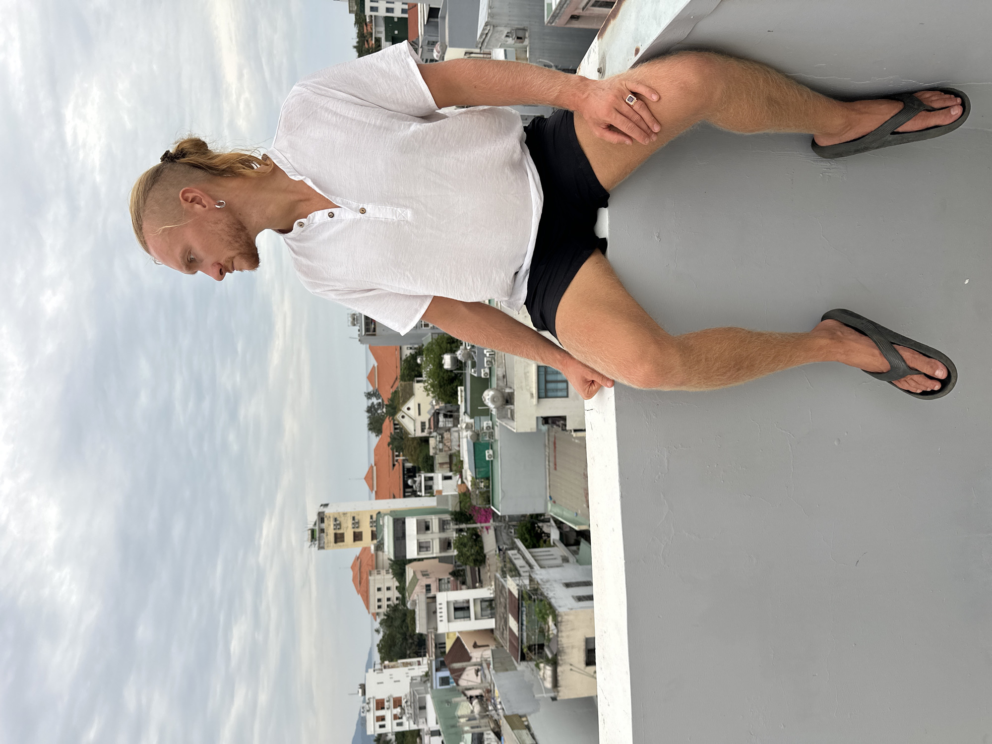 A fair-skinned man with blonde hair pulled back in a bun and a short beard sits on a grey concrete ledge overlooking a dense city skyline. He is wearing a loose white short-sleeved shirt with a Henley neckline and black shorts. His legs are bare and hairy. He wears dark grey flip-flops on his feet and a large ring on his right hand. The city below consists of a mix of residential and commercial buildings, many with orange tiled roofs, and some taller, more modern structures. The sky is overcast with grey clouds. The ocean is visible in the distance behind the buildings. The scene appears to be in a coastal city in Vietnam, likely Đà Nẵng, given the architectural style of the buildings and the topography.