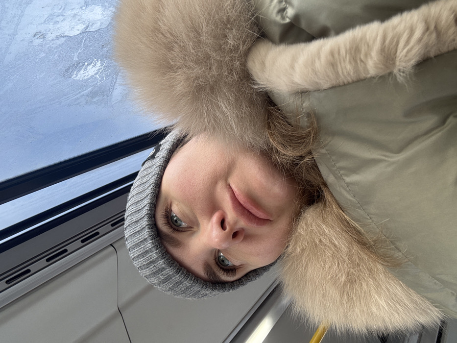 A person wearing a gray knit hat and a large fur-trimmed coat looks toward the camera. The individual's eyes are a striking green or gray. A window showing a cloudy sky is visible in the top left of the frame, suggesting an indoor setting. The perspective is slightly from below, looking up at the person. The image is rotated, so the person's head appears to be tilted.