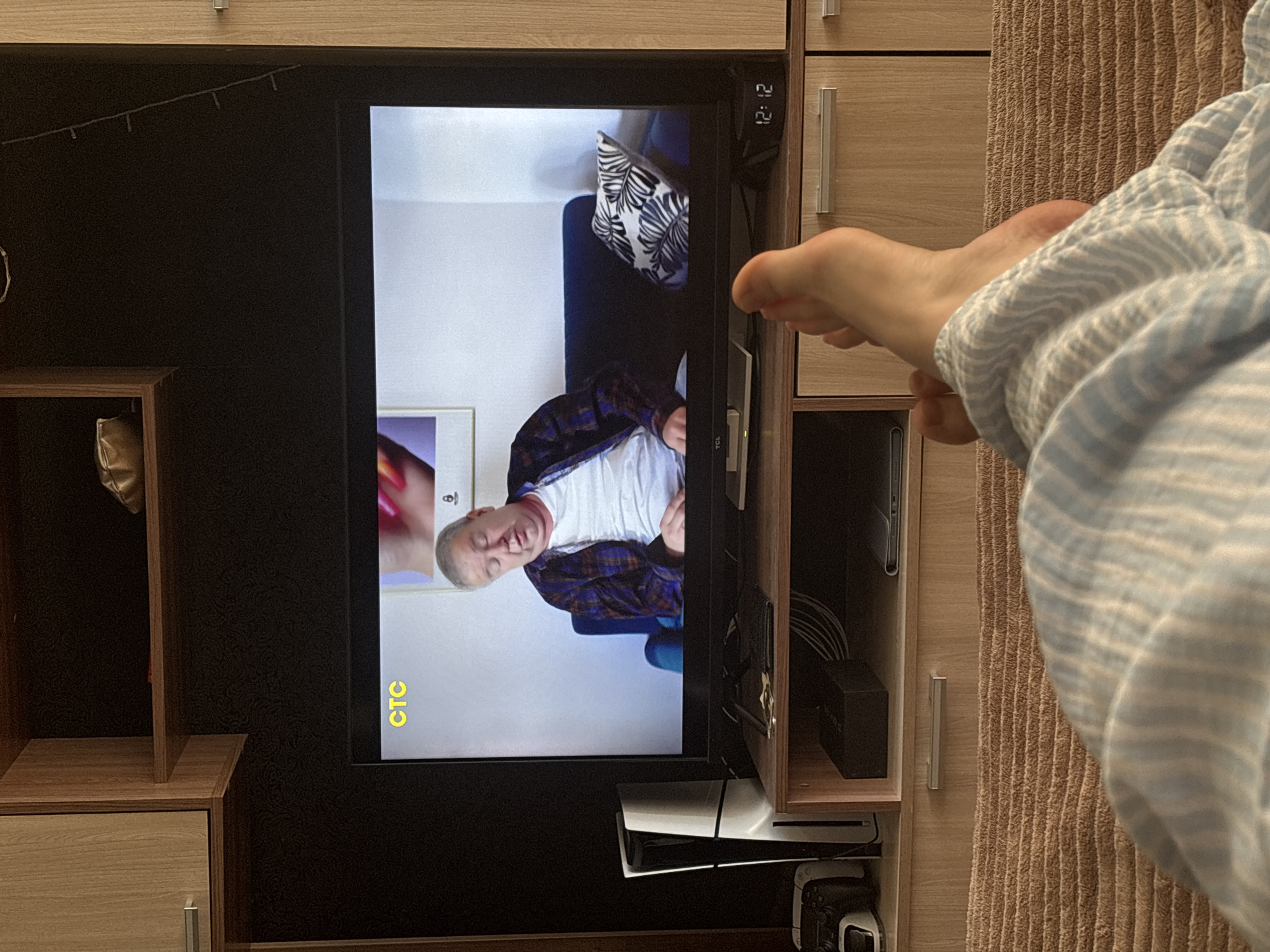 A television displays a man on screen, positioned within a wooden entertainment unit. Below the television, a Playstation 5 console and a game controller are visible. The foreground features a person covered with a striped blanket. The location of this image is not discernible from the provided details.