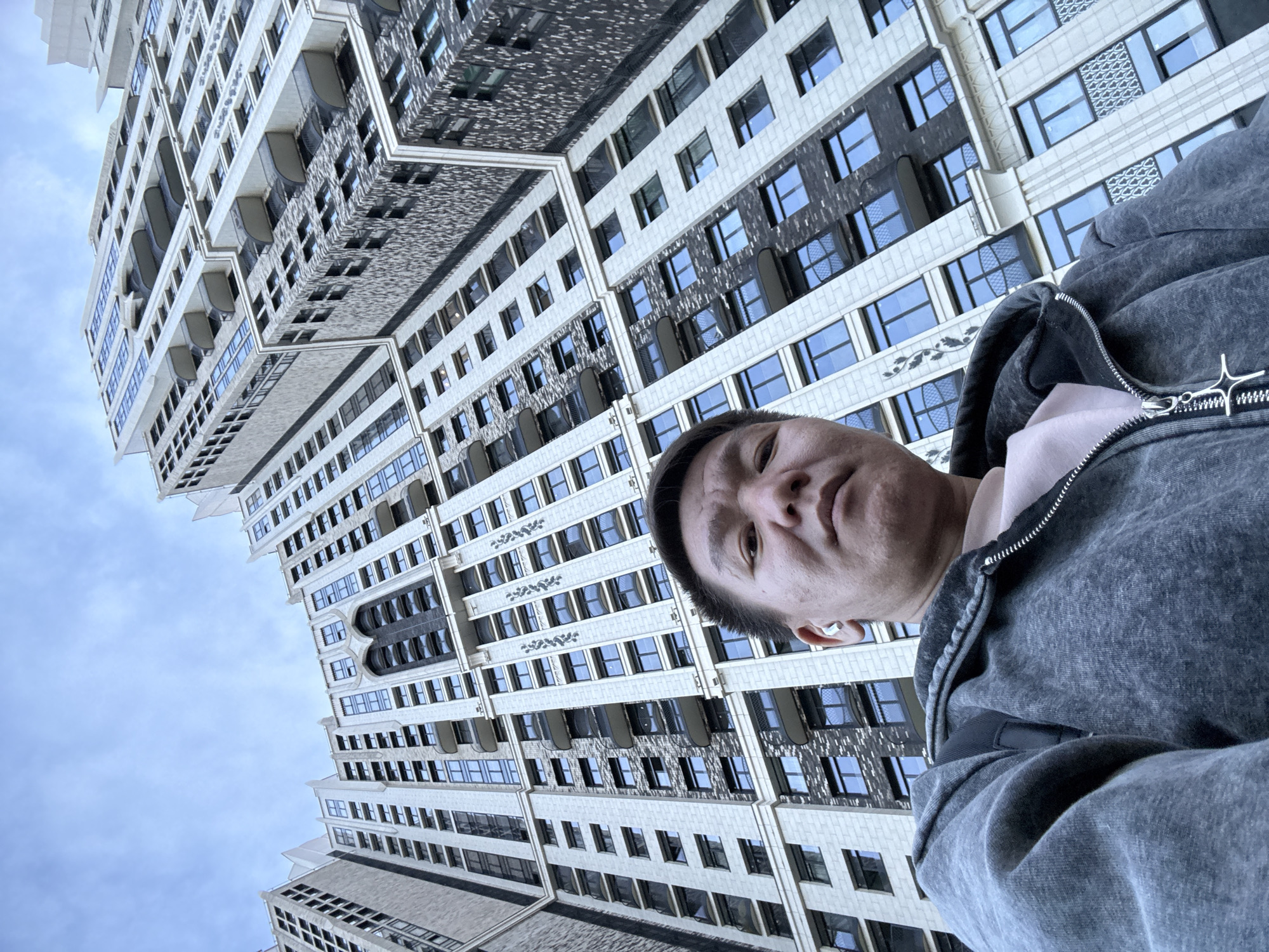 A low-angle selfie featuring a person with short dark hair, wearing a gray hooded sweatshirt over a light pink top, with a white wireless earbud visible in their left ear. The person is looking directly at the camera. The background is a tall building in Khimki, Russia, with multiple floors and windows. The building's facade is predominantly white, with some sections featuring darker gray or black panels. The sky is visible in the upper left corner.