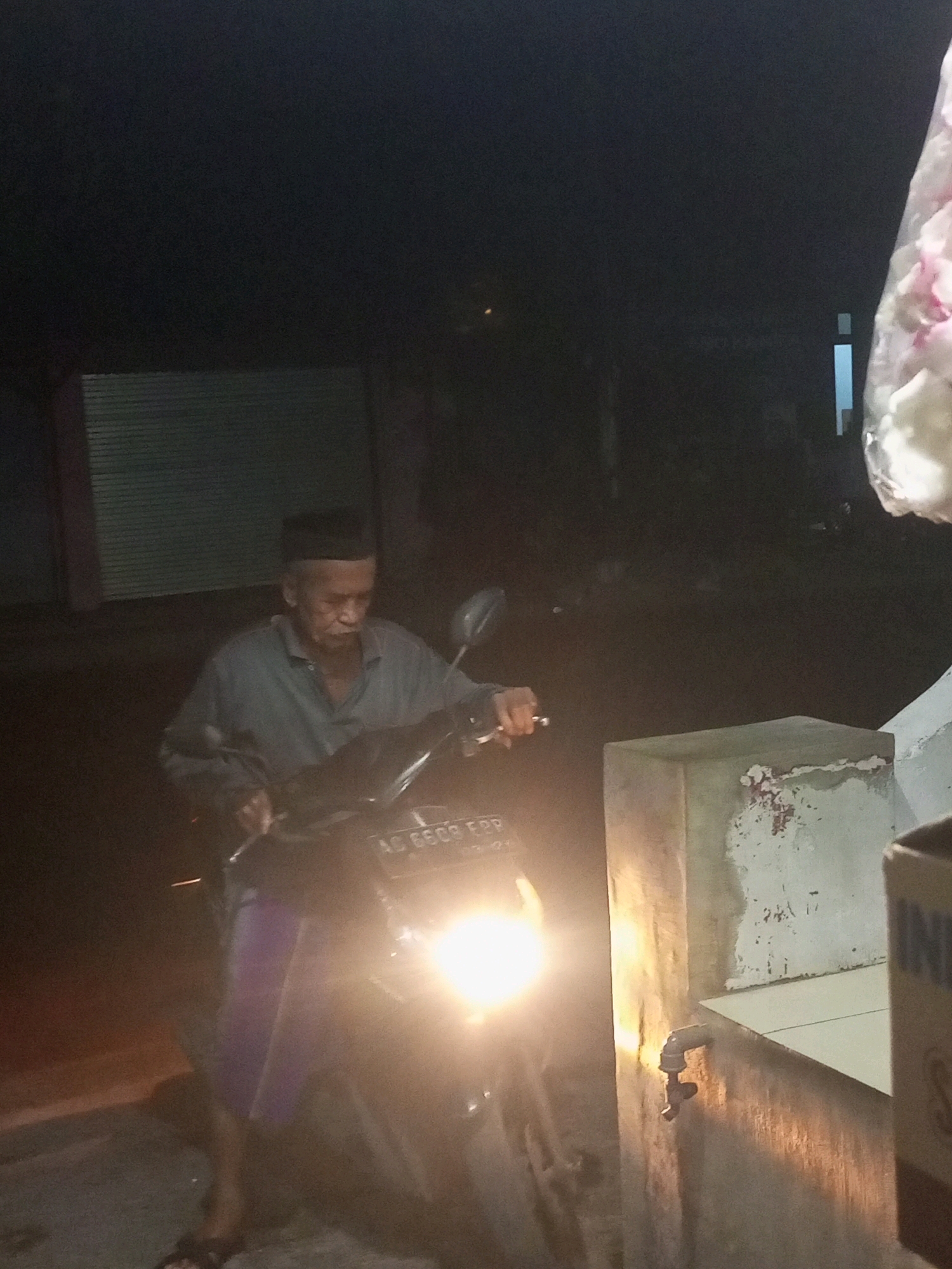 In the city of Ngasem, Indonesia, at nighttime, an elderly man wearing a black peci hat and a grey shirt is seated on a dark-colored motorcycle. The motorcycle's headlight is illuminated, casting a bright glow on the scene. The man's facial features are visible, with his mouth slightly open. He appears to be holding the motorcycle's handlebars. The background is dark and out of focus, with some faint vertical lines suggesting a building or structure. To the right, a concrete pillar with a worn, textured surface stands, and a metal faucet is attached to it. Part of a white and pink object, possibly flowers wrapped in plastic, is visible in the upper right corner of the frame. The ground is dark and appears to be asphalt.