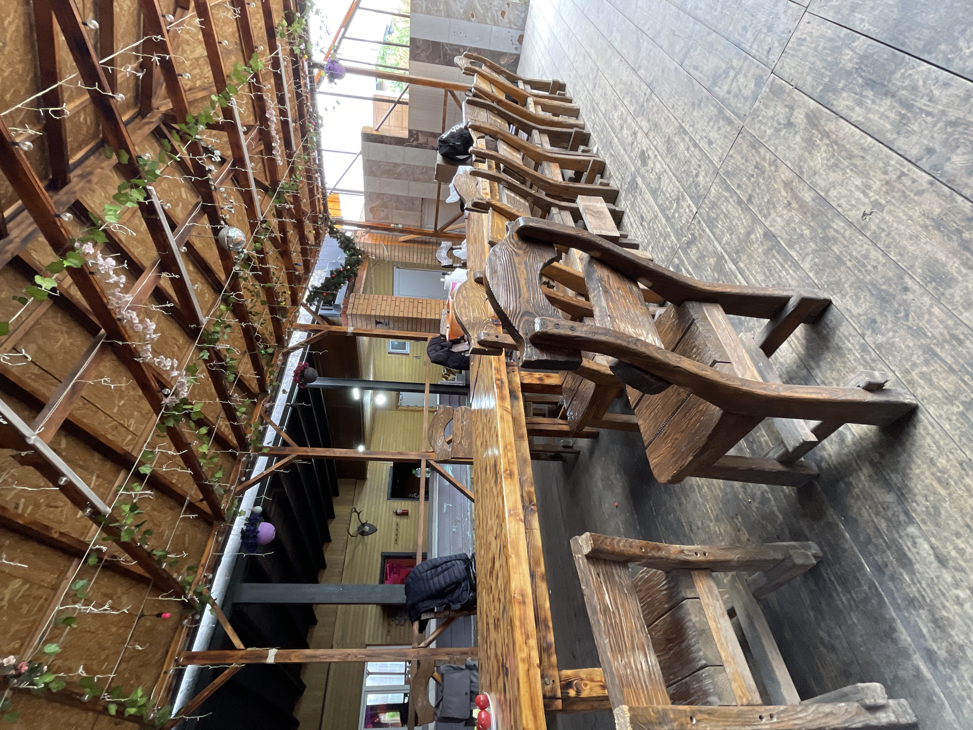 The image depicts a long wooden table with attached wooden chairs, likely in a restaurant or outdoor seating area in Картмазово, Russia. The table and chairs appear rustic and handcrafted. Above the table, there is a wooden structure with string lights and decorative garlands. The flooring is made of wood planks. Some personal belongings like bags are placed on the chairs. The background reveals the interior of a building with windows and other architectural features.