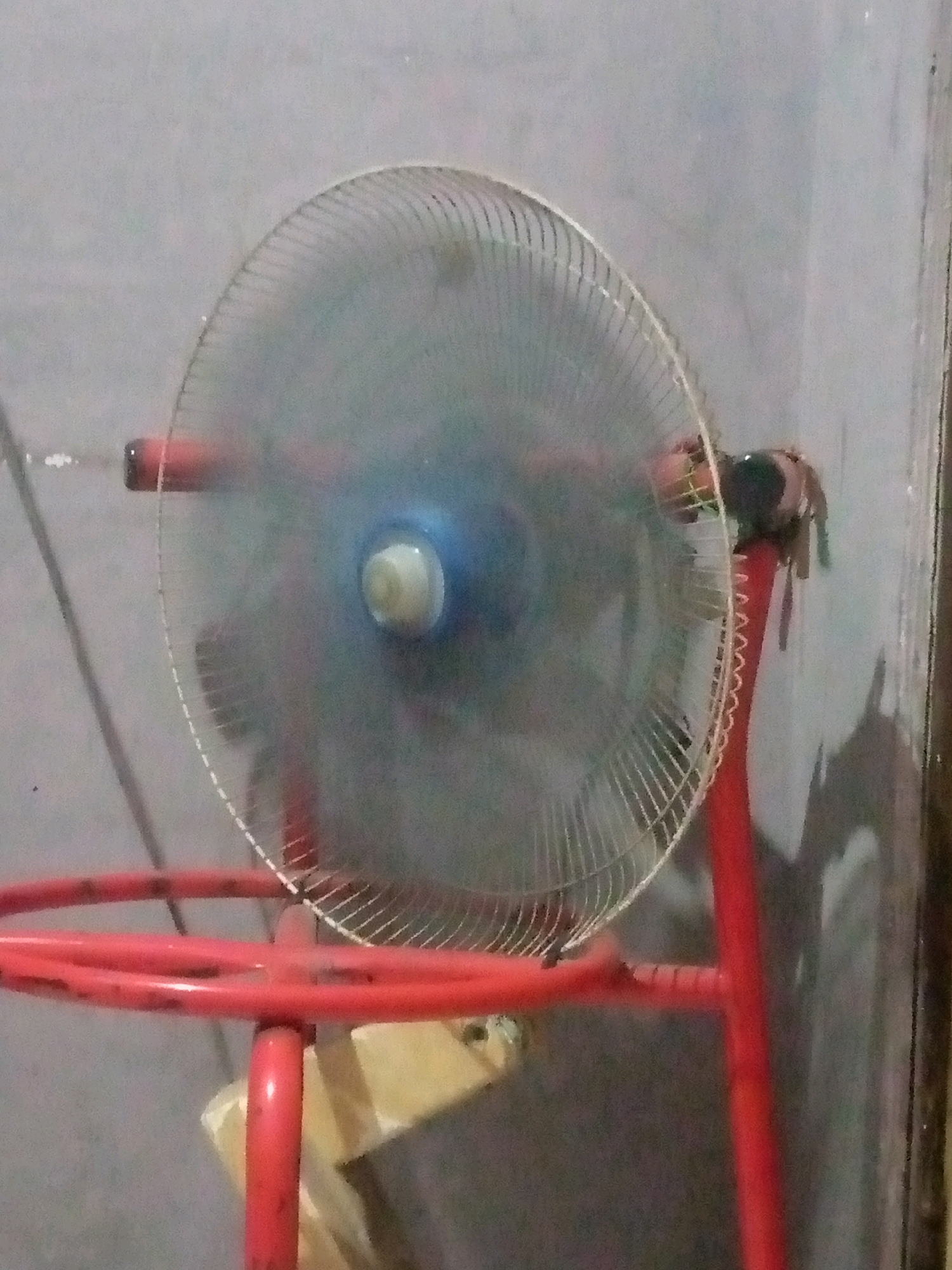 An out-of-focus, close-up shot captures a pedestal fan. The fan's grill is visible with thin wires, and the central blue hub with a white knob is discernible. The fan is mounted on a bright red metal stand. The background consists of a textured, light-colored wall and a darker, vertical surface on the right. The overall lighting is dim, creating soft shadows on the wall behind the fan. The image quality is poor, with significant blur throughout.