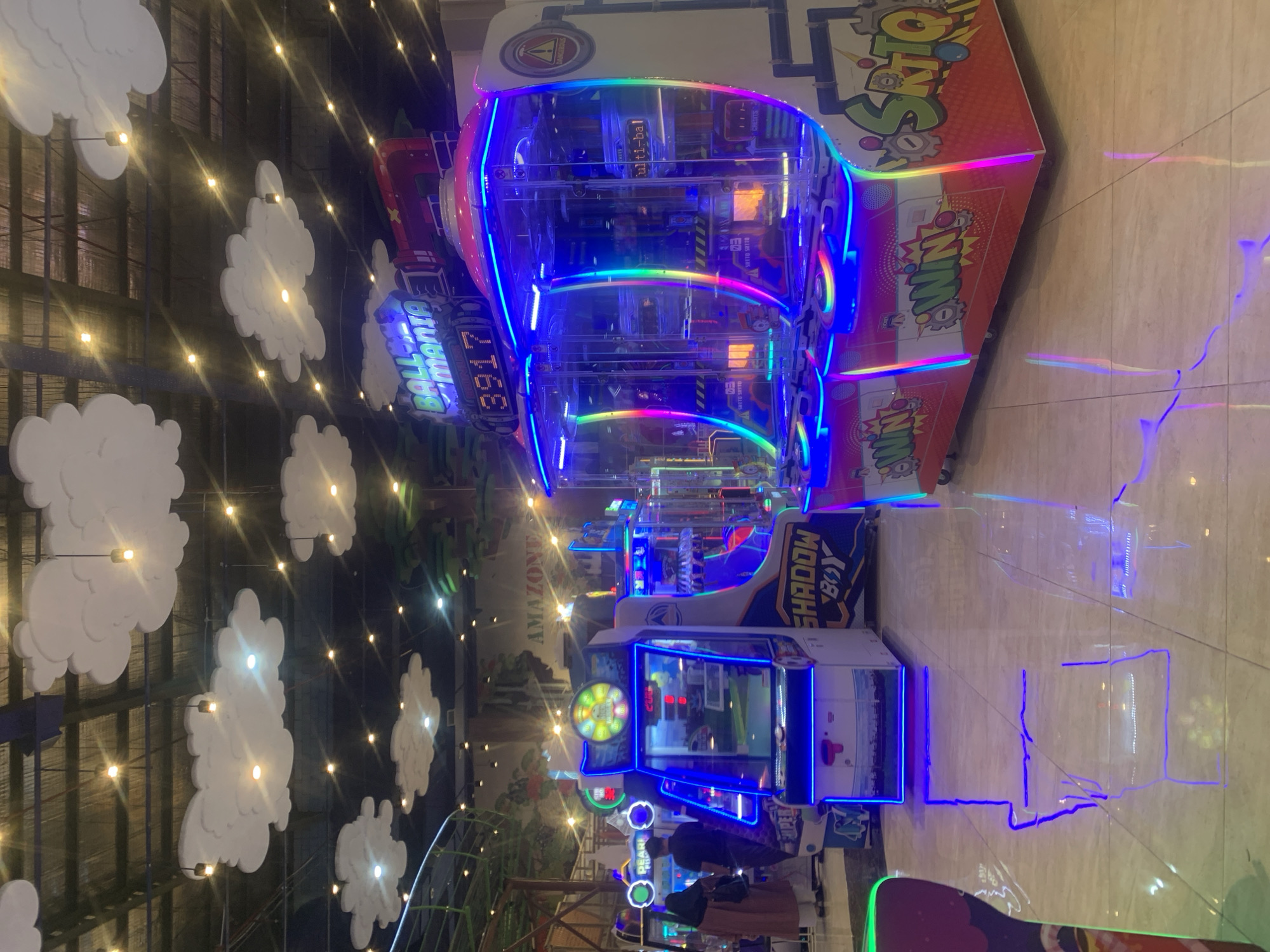 This image depicts a lively indoor entertainment center, likely an arcade or family fun zone, in Samarinda, Indonesia, illuminated by numerous vibrant lights, suggesting it's nighttime or a low-light setting. The scene is dominated by several brightly lit arcade machines with colorful LED accents.
In the center, a large claw or prize redemption game is prominent, featuring a clear casing and a cascade of rainbow-colored and blue LED lights. On this machine, the words "BALL MANIA" and the number "3917" are visible, along with "ulti-bal" and "CREDITS." Another adjacent machine prominently displays "WIN."
Further into the background, other arcade games can be seen, including one labeled "SHADOW BOY" and "AMAZONE," glowing with blue lights. A few indistinct figures of people are visible in the background, appearing to be enjoying the games, with one person in a brown skirt near a machine displaying "PEARL."
The walls are decorated with multiple white, cloud-shaped cutouts, illuminated by warm, glowing string lights, creating a whimsical atmosphere. The floor is a reflective, light-colored tiled surface, mirroring the colorful lights from the arcade machines in long streaks. Additional visible text includes "SRITO SRITO." The overall impression is one of fun and energetic entertainment.