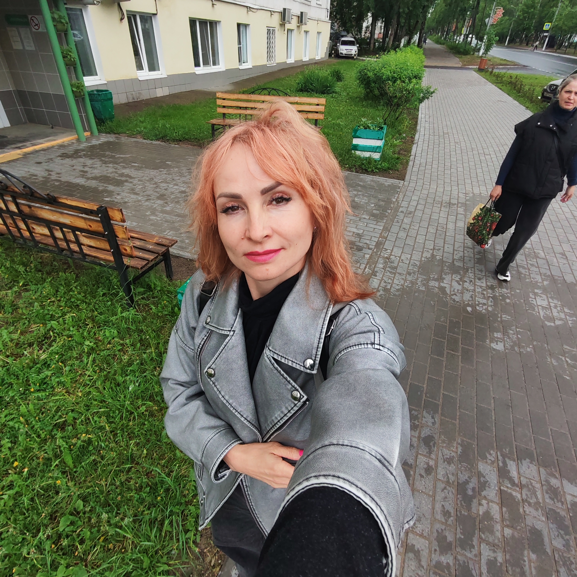 The image is a selfie taken outdoors, seemingly on a cloudy or overcast day in the city of Chaikovsky, Russia. The person taking the selfie is a woman with reddish-blonde hair, wearing a gray leather jacket over a black top. She has on makeup.
In the background, there is a wet paved sidewalk leading past a grassy area with benches towards a street. Another woman is walking along the sidewalk, wearing a black vest, dark pants, and carrying a shopping bag. A multi-story building with light yellow siding and white-framed windows is also visible. Trees line the street.