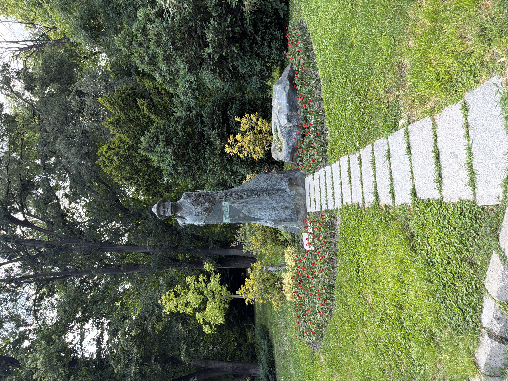 This image captures a bronze statue of a man with a prominent mustache and hat, situated in a well-maintained park environment. The statue, depicting A. Bozhinov, stands on a textured stone base, with the inscription "А. БОЖИНОВ" (A. BOZHINOV) visible on its right side. The man is depicted wearing a long coat and appears to hold a decorative element or staff in his left arm.
A light gray stepping stone path leads directly towards the statue. Flanking the path and the statue are two curved flower beds filled with numerous small red flowers, likely Salvia splendens. Behind these flower beds, various green and yellow-leaved shrubs provide additional landscaping. To the immediate right of the statue, within the flowerbed arrangement, lies a large, smooth, dark gray rock with a small yellow-leaved plant growing from its surface.
The ground cover consists of a mix of manicured green grass and clover-like plants. In the midground and background, a dense assortment of mature trees with varying foliage shades and heights indicates a lush park setting. The sky is partially visible through the tree canopy in some areas, suggesting an outdoor daytime scene.
This scene is located within Tsar Simeon's Garden in Plovdiv, Bulgaria, where the monument to Alexander Bozhinov (A. Bozhinov), a renowned Bulgarian artist and writer, is prominently displayed.