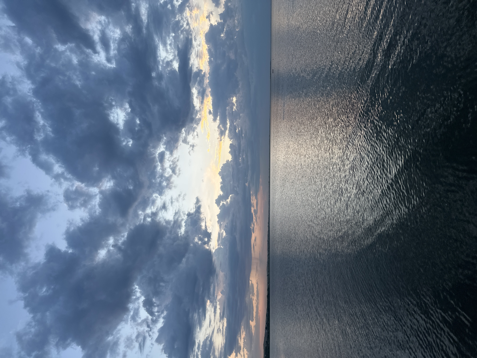 A serene body of water stretches towards the horizon under a dramatic sky filled with heavy, dark clouds. The water's surface is gently rippled, reflecting the muted light of what appears to be dusk. In the distance, a faint line of land is visible, suggesting the presence of a coastline or distant hills. The clouds are textured and varied, with lighter patches of sky peeking through, hinting at the lingering light of sunset. The scene evokes a sense of calm and natural beauty. The location is specified as the city of Kokshetau, Kazakhstan.