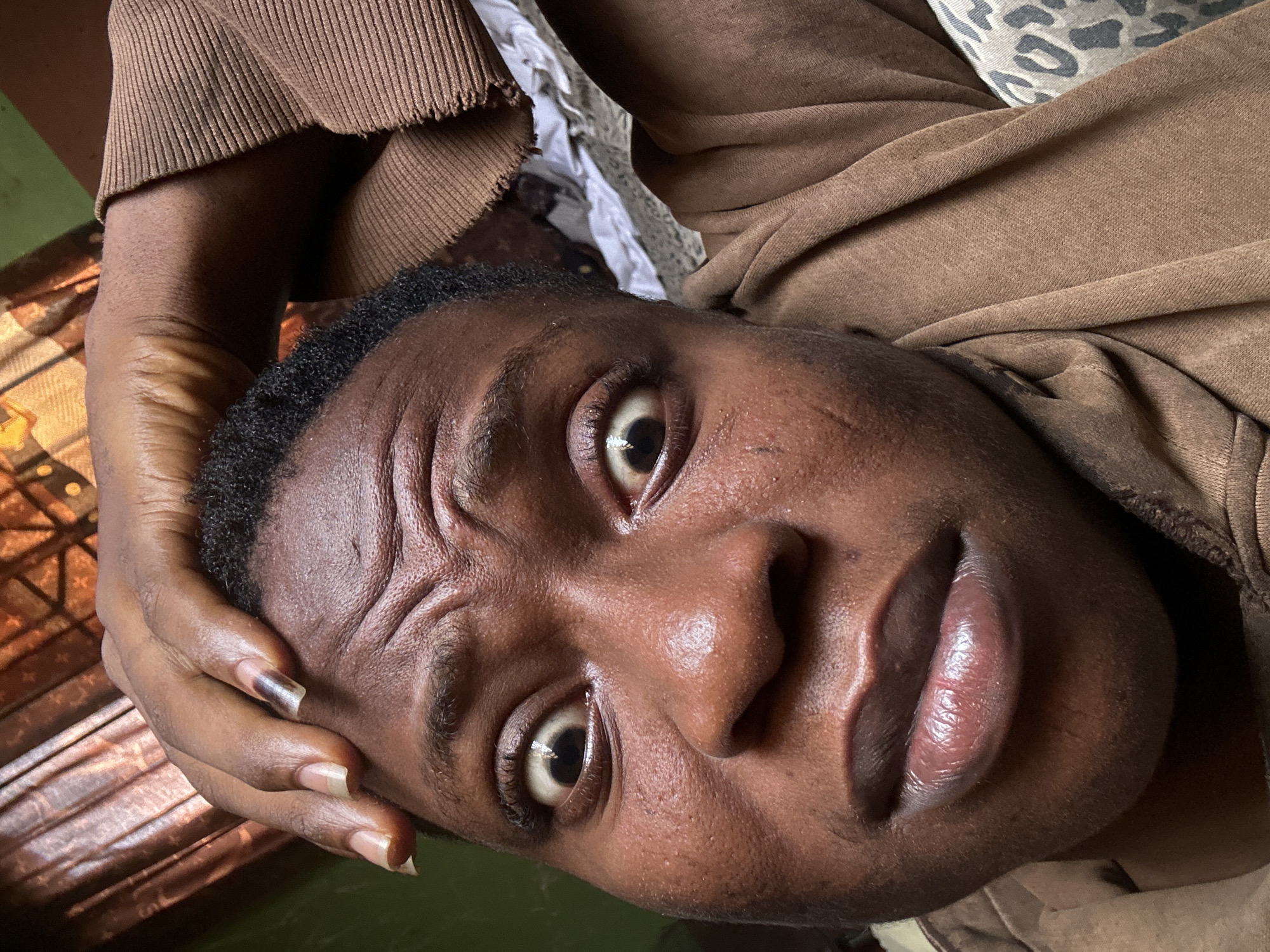 A close-up, angled view of a dark-skinned person. They are wearing a brown, long-sleeved garment. Their right hand is near the side of their face. Visible features include dark eyes and full lips. The environment appears to be indoors, with a window and curtain visible in the background. There is no visible indication of the location being Ikorodu, Nigeria.