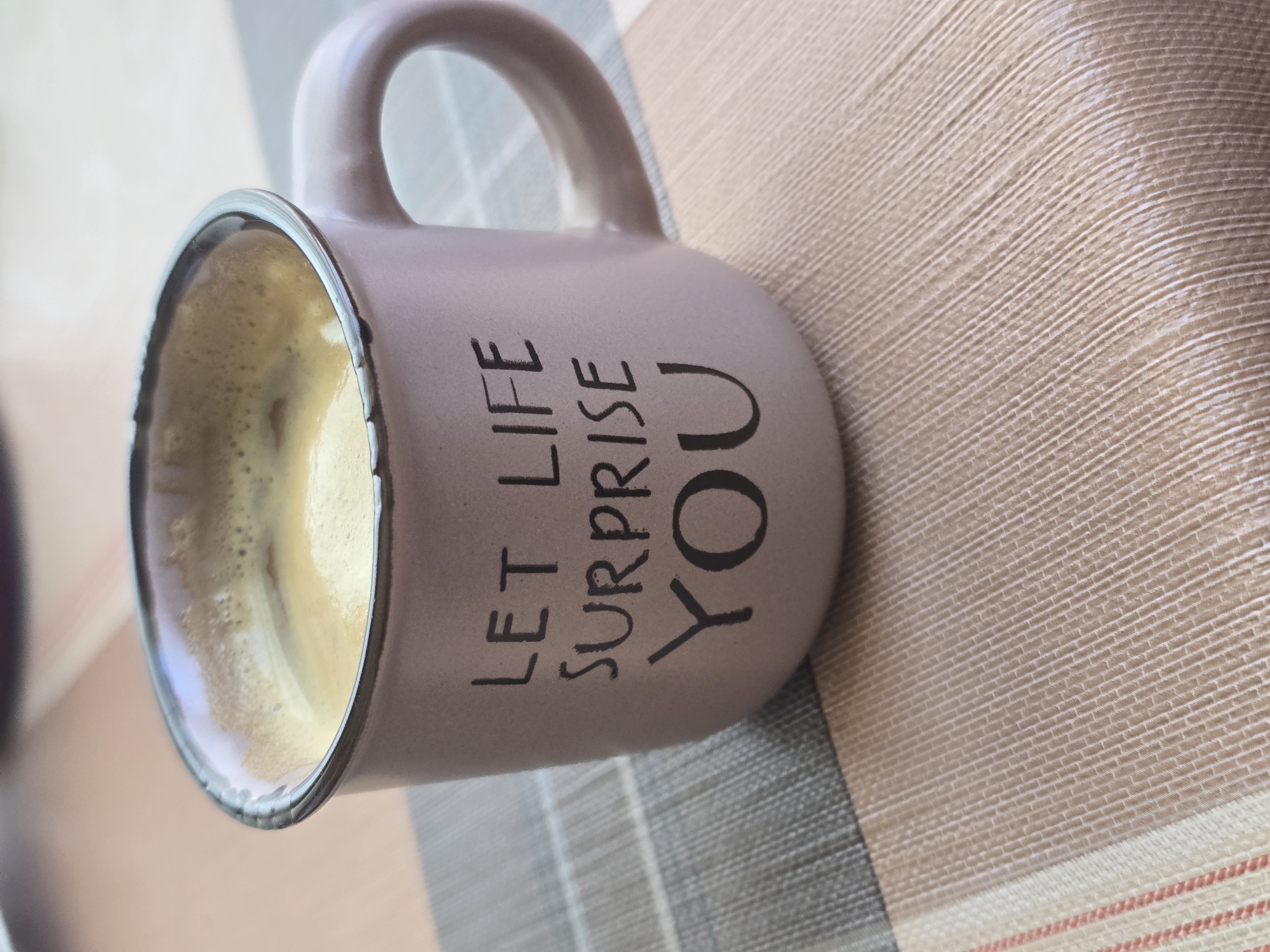 A close-up shot shows a light purple mug with the phrase "LET LIFE SURPRISE YOU" printed in black letters. The mug is partially filled with a light brown liquid, likely coffee or tea. It rests on a textured surface that appears to be a tablecloth or placemat with a striped pattern in neutral tones. The scene is not identifiable as being specifically located in Pernik, Bulgaria, as the image provides no contextual clues.