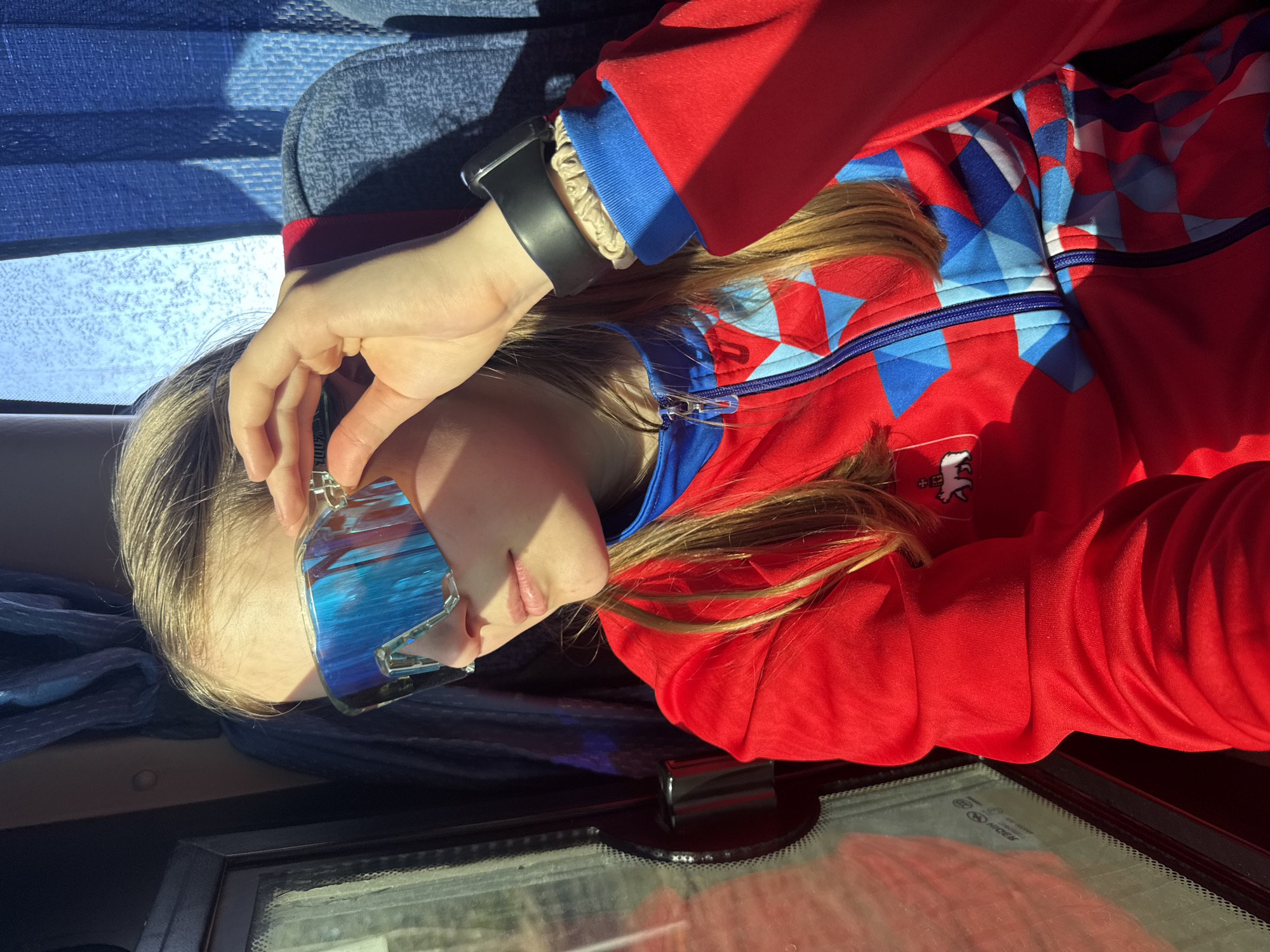 A young person with blond hair is resting in a seated position, possibly in a vehicle. They are wearing a red and blue patterned jacket and mirrored sunglasses. The person has a black smartwatch on their left wrist. The location is unclear, and there is no definitive evidence to confirm the scene is in the city of Челва, Russia. There are reflections in the window of what appears to be some vegetation.