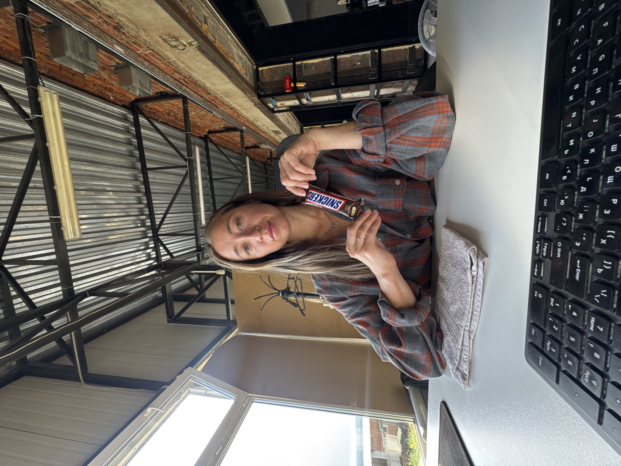 Here is a detailed description of the image:
**Scene:** The photo appears to be taken indoors, likely in a non-traditional office setting, possibly a renovated industrial space or a home office with an industrial aesthetic.
**People:** A woman is visible. She is holding a Snickers bar.
**Objects:**
* A Snickers bar
* A keyboard
* A gray towel
* A microphone stand
* A window
* A mousepad
* Office chair
* Fluorescent lights
* Metal roofing structures
**Environment:** The background shows a distinctive architecture with metal roofing and beams, suggesting the industrial design. A window provides some natural light.
**Location:** There is no direct visual information in the image to conclusively confirm that it is located in Chaikovsky, Russia. Therefore, that cannot be determined.