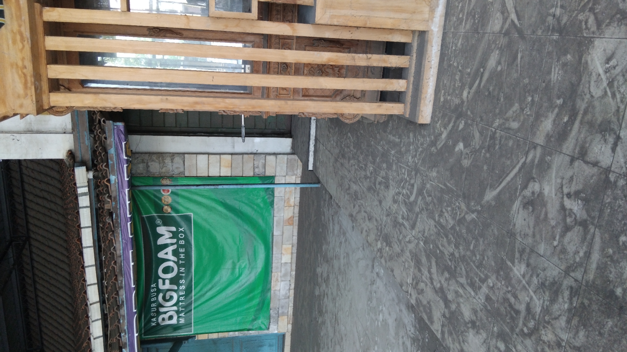 The image shows a storefront in Batang, Indonesia, featuring a prominent green banner with white text that reads "KASUR BUSA BIGFOAM MATTRESS IN THE BOX". To the right of the banner is a wooden structure with vertical slats, possibly part of a display or entrance. The storefront is situated on a paved outdoor area. The building behind the banner appears to have a tiled roof and light-colored walls. The overall scene suggests a commercial establishment.
