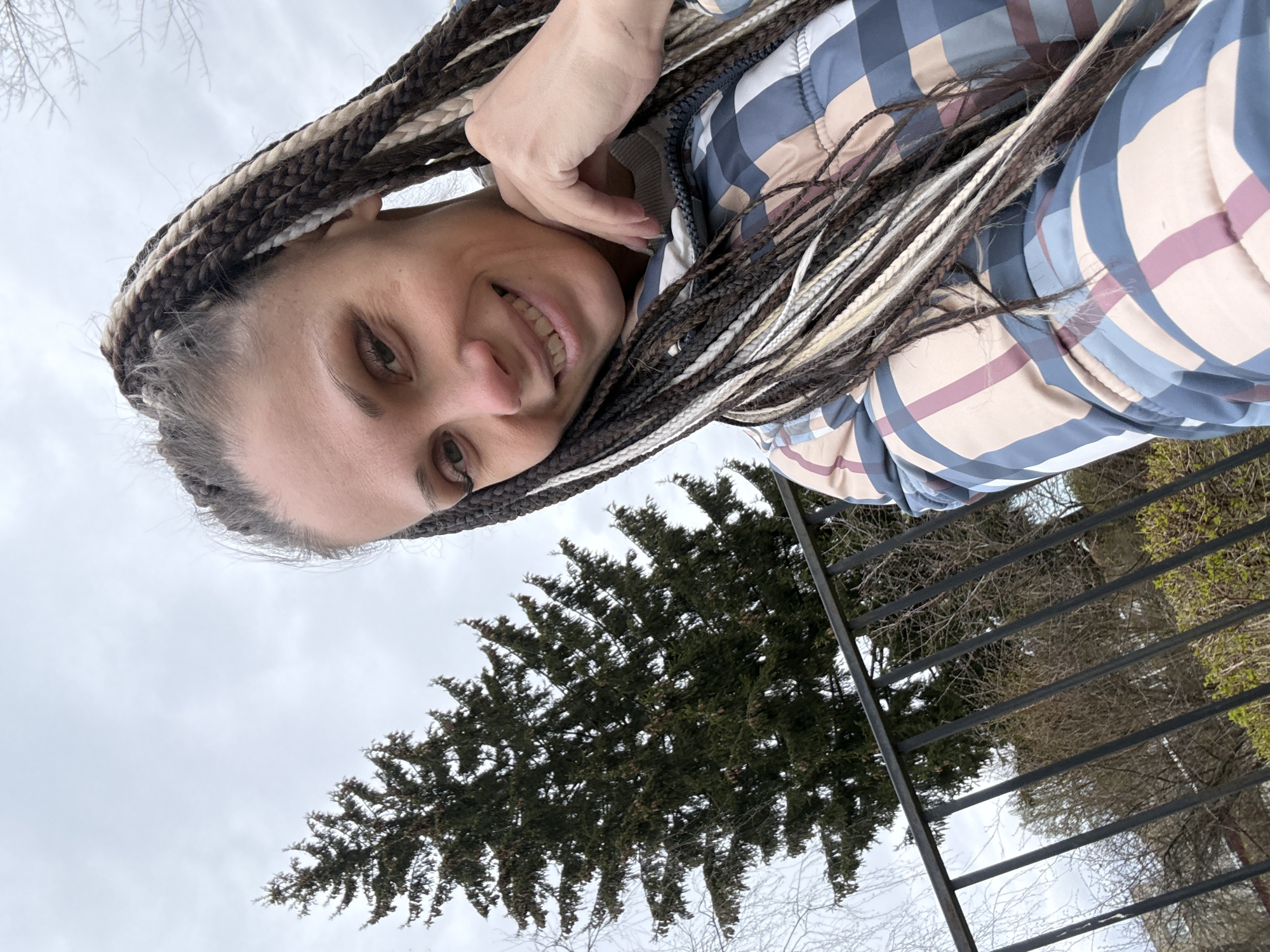 A woman is featured in a close-up selfie, captured from a low, upward-looking angle, under an overcast sky in Rusanovo, Russia. She is smiling broadly at the camera, her expression friendly and engaging. Her hair is styled in long, multi-toned box braids, showcasing shades of dark brown, blonde, and white or grey. She wears a padded jacket with a distinct plaid pattern in blue, light brown, maroon, and white. One of her hands is visible near her head.
The background reveals an outdoor setting. A prominent, dark green evergreen tree with dense foliage stands behind a dark metal fence composed of horizontal bars. Beyond these, hints of bare deciduous trees are visible against the cloudy, light grey sky, suggesting a cooler time of year. The image is rotated approximately 90 degrees counter-clockwise, creating an unusual orientation. No text is visible in the media file.