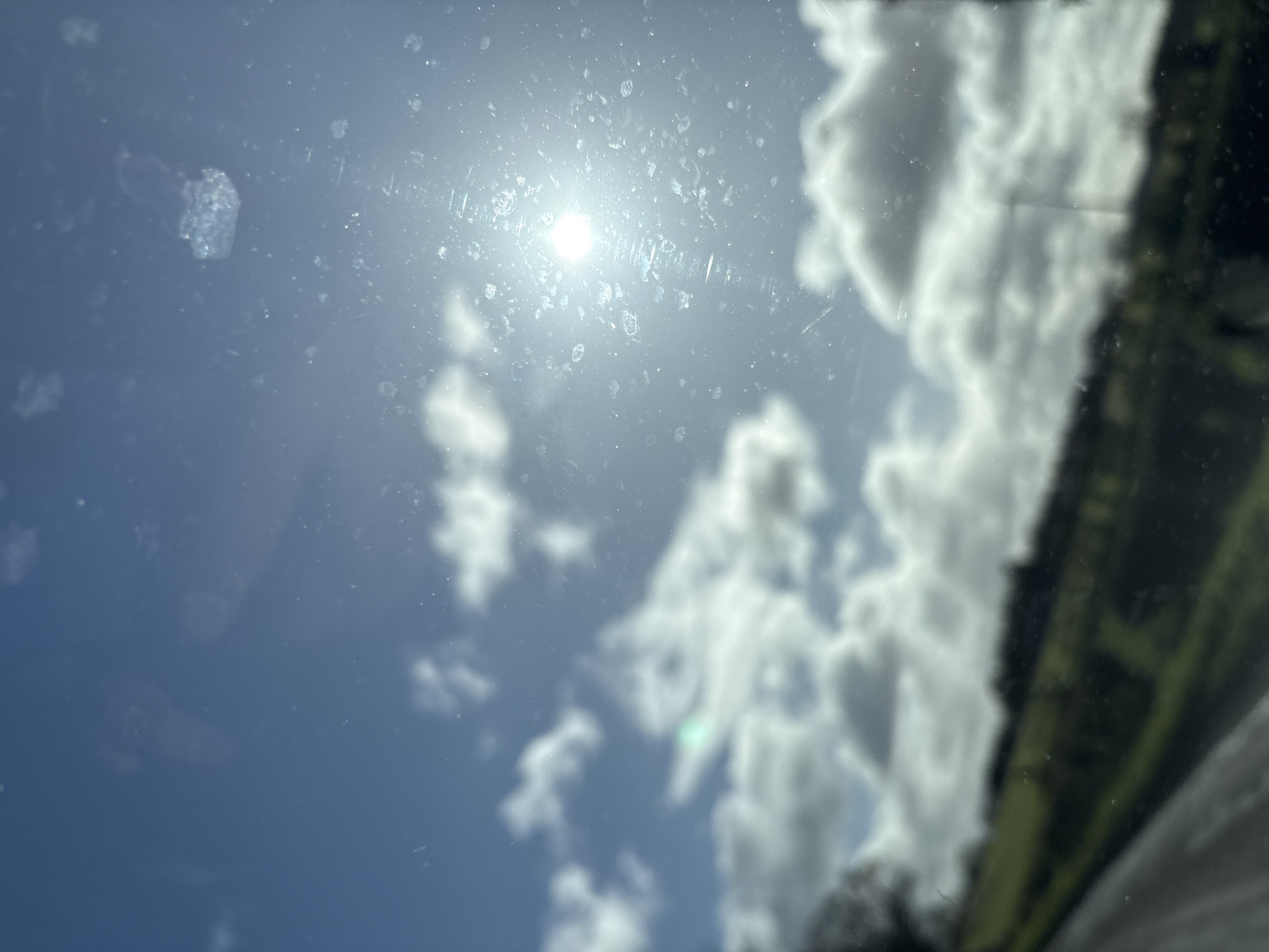 The image depicts an outdoor daytime scene captured through a significantly smudged and dusty transparent pane, likely a vehicle window. The location is Ruyton-XI-Towns, United Kingdom. The upper two-thirds of the frame show a blue sky with scattered white clouds. The sun, a bright spherical light source, is positioned in the upper right quadrant, creating lens flare and illuminating numerous sharp, irregular smudges, dust particles, and streaks on the glass surface. The lower third of the image reveals a highly blurred rural landscape, consisting of green fields or hills sloping upwards from left to right, interspersed with indistinct dark vegetation such as trees or hedges. A dark, diagonal element is visible along the bottom left edge, consistent with a road surface or vehicle interior component. No individuals are present, and no discernible actions or interactions occur within the external environment.
