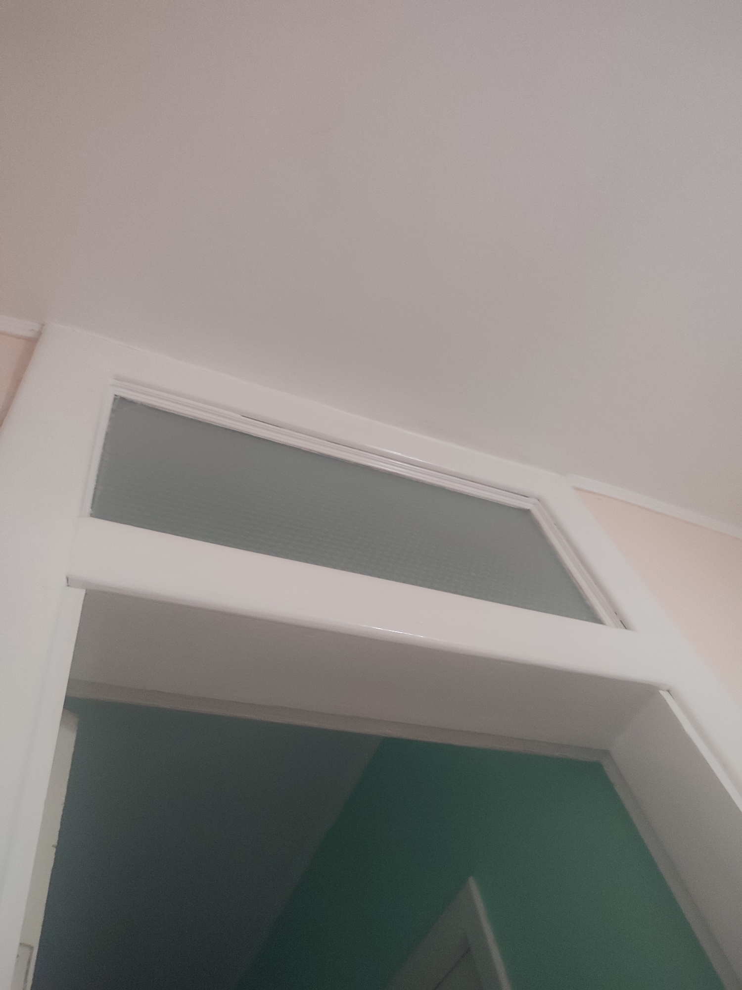 The image shows a close-up, low-angle view of a doorway with a transom window. The doorway is framed in white, with a frosted glass pane in the transom. The wall to the left of the doorway is painted a pale pink, and the wall below the transom, visible through the open doorway, is painted a teal green. The ceiling is a neutral, light color. The image is taken indoors, suggesting it could be part of a residential or commercial building. There are no people or animals visible. The scene is static and offers no indication of any ongoing activity.