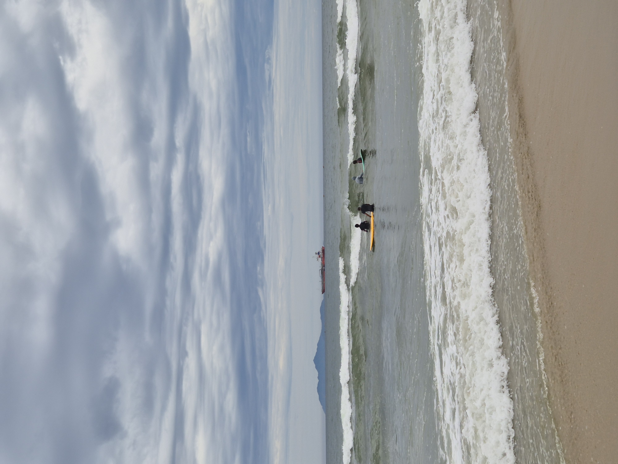 The image shows a cloudy sky over the ocean with a beach in the foreground. In the middle distance, there are three people in the water, two on surfboards and one kneeling. A large cargo ship and a distant island are visible on the horizon. The scene is likely of My Khe Beach in Da Nang, Vietnam, known for its surfing and beautiful coastline.