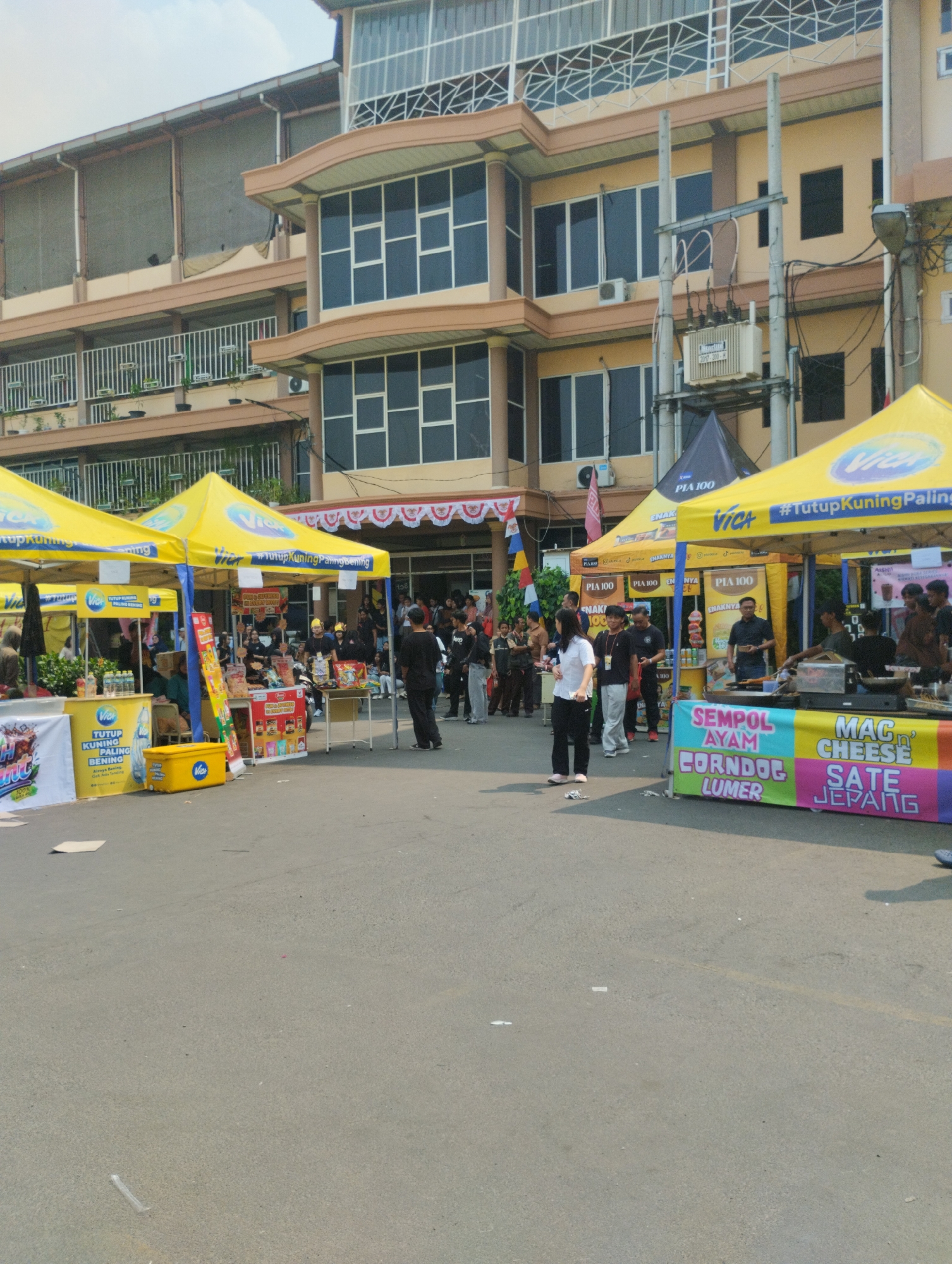 The image depicts an outdoor market scene in front of a large, multi-story building in Bekasi, Indonesia. Yellow tents, branded with "Vica" and "#TutupKuningPalingBening", are set up along a paved area, suggesting a commercial event or bazaar. These tents house various food stalls, indicated by banners displaying menu items such as "Sumpol Ayam Corndog Lumer" and "Mac n Cheese Sate Jepang". Several people are present, some browsing the stalls, others working behind them. A few individuals are interacting, while others stand individually. The building behind the stalls is light yellow with numerous windows and a modern architectural style. Utility poles with electrical equipment are visible in the background. The ground is asphalt, with some litter scattered on it. The lighting suggests it is daytime, likely morning or afternoon.