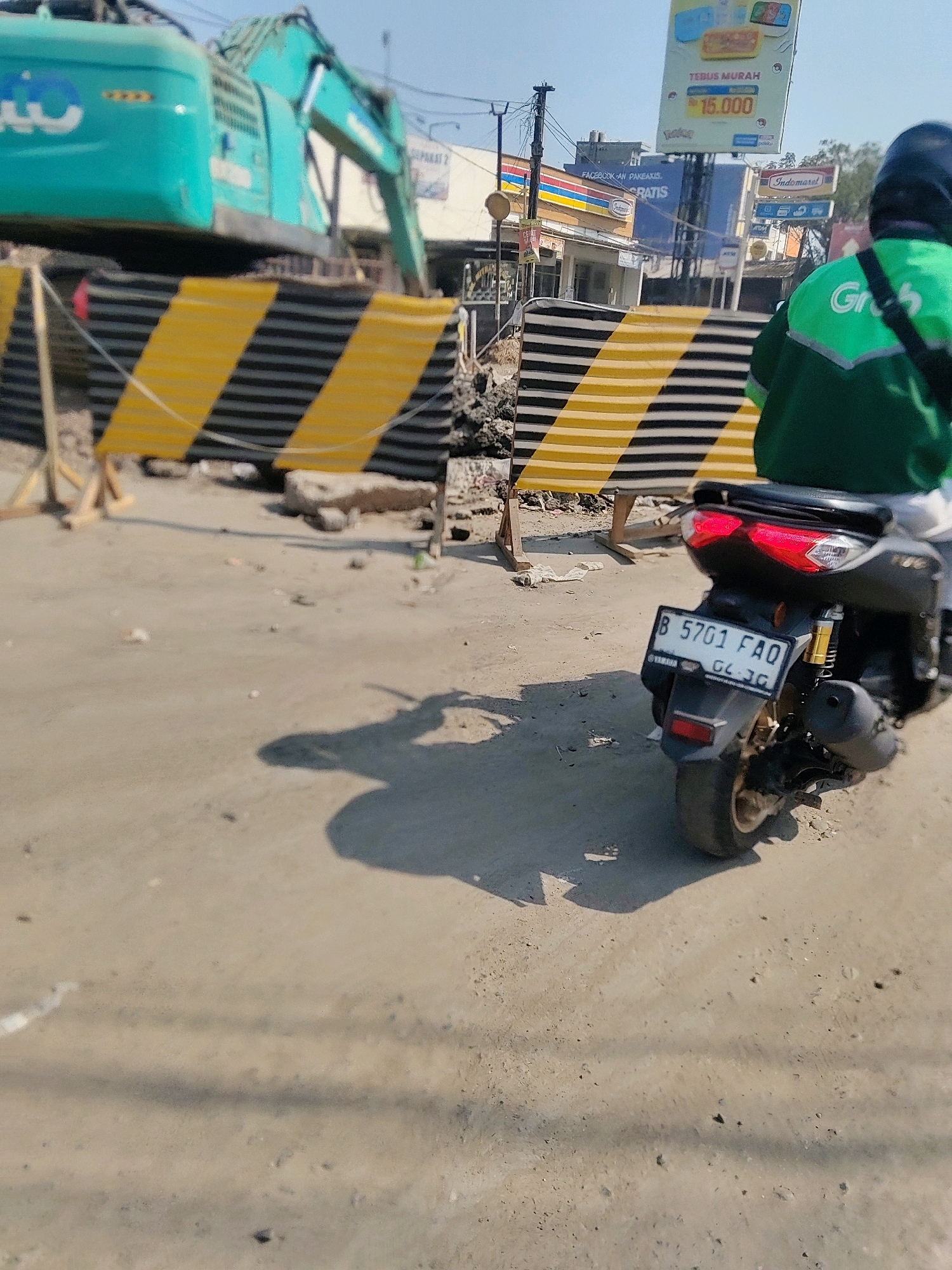 In Pasirsari, Indonesia, a construction site is evident with a large green excavator. A black and yellow striped barrier separates the construction area from a dirt road. A person on a black motorcycle, wearing a green jacket with the "Grab" logo, is stopped on the road, facing away from the camera. The motorcycle's license plate reads "B 5701 FAQ". Buildings, including what appears to be an Indomaret store and other commercial establishments, are visible in the background. The sky is clear and blue, indicating a sunny day.