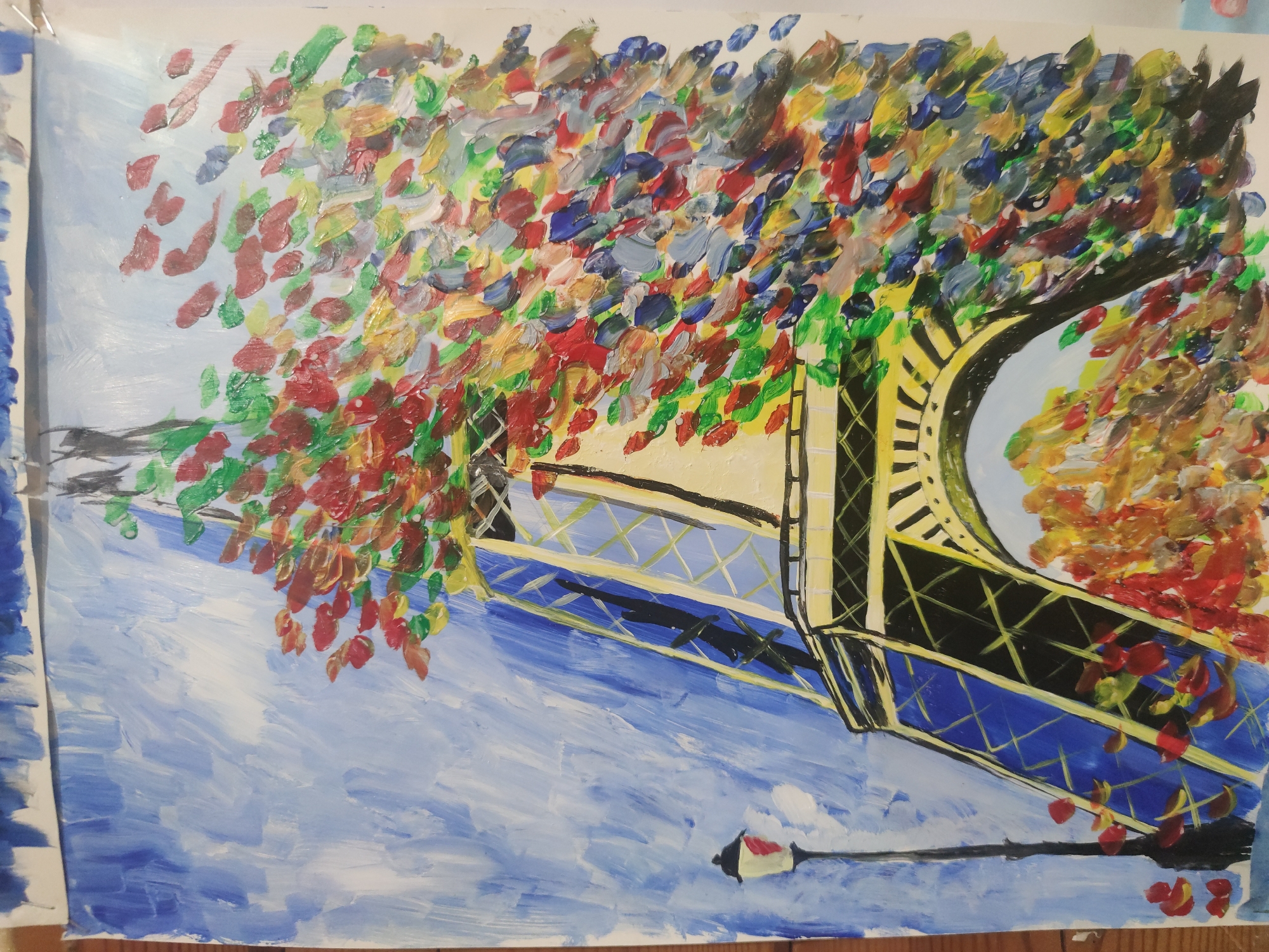 This is an impressionistic painting of a bridge, likely the Eiffel Tower, seen from below. The bridge is depicted in dark black and yellow with crisscrossing lines, suggesting its intricate metal structure. Above and around the bridge, the leaves of a tree are painted in a vibrant, abstract style with dabs of red, green, yellow, and blue, hinting at autumn foliage or a colorful flowering tree. The sky is a soft, hazy blue with white streaks, possibly indicating a cloudy or windy day. Below the bridge, the scene transitions to a body of water, rendered in broad strokes of light and dark blue, giving the impression of gentle ripples. A single lamppost with a red top is visible on the right side of the painting, adding a touch of quaintness to the scene. The overall impression is one of a bustling, colorful, and dynamic urban landscape, possibly during a pleasant season.