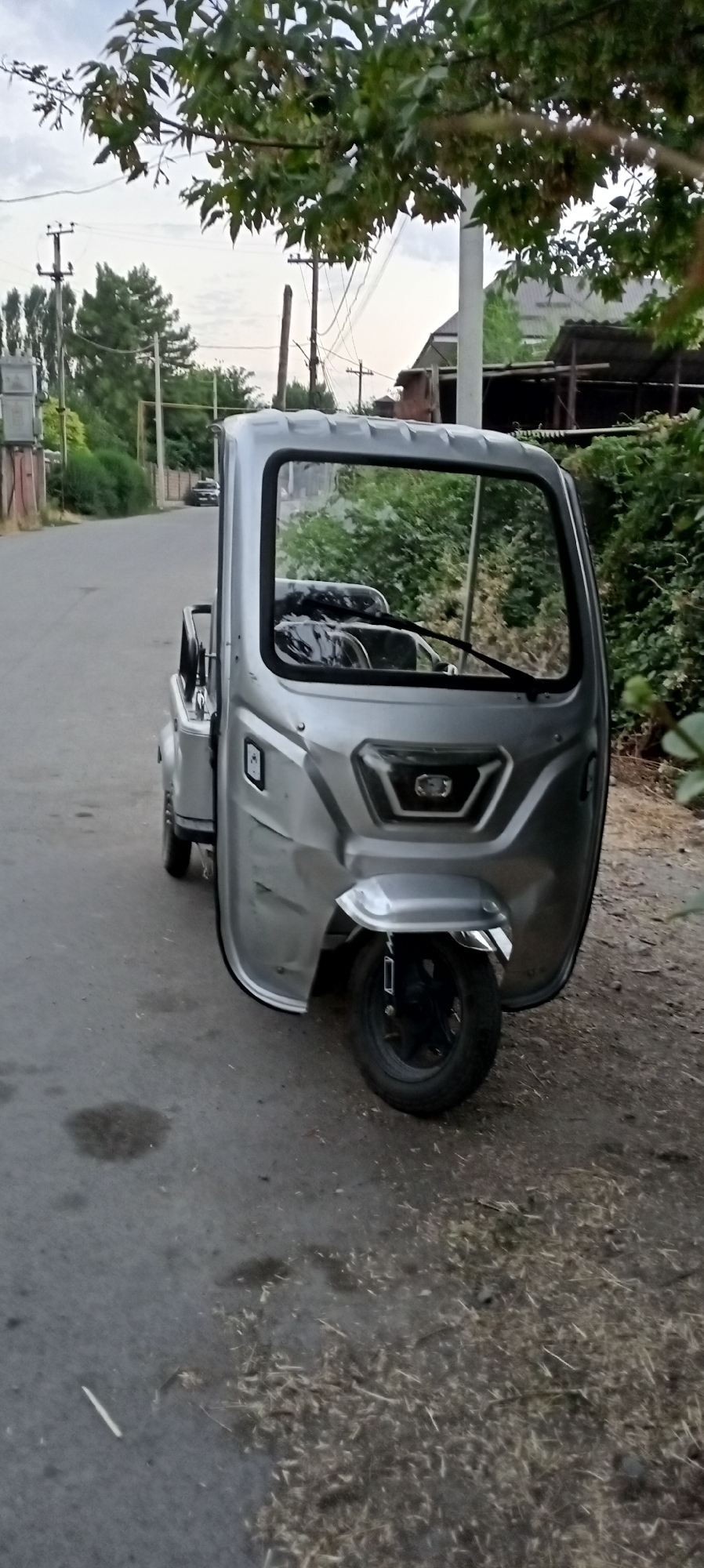 A three-wheeled, silver motor vehicle is parked on the side of a paved road. The vehicle has a covered cab with a windshield and a single wheel in the front. The front of the vehicle has a chrome fender over the front wheel. The rear of the vehicle appears to be a cargo bed. The vehicle is parked on the right side of the road, facing away from the camera. The road is an asphalt surface and there is some debris, including dry grass and twigs, on the shoulder of the road. In the background, there are trees, utility poles with wires, and buildings. The sky is overcast. This scene is located in the city of Sokuluk, Kyrgyzstan.