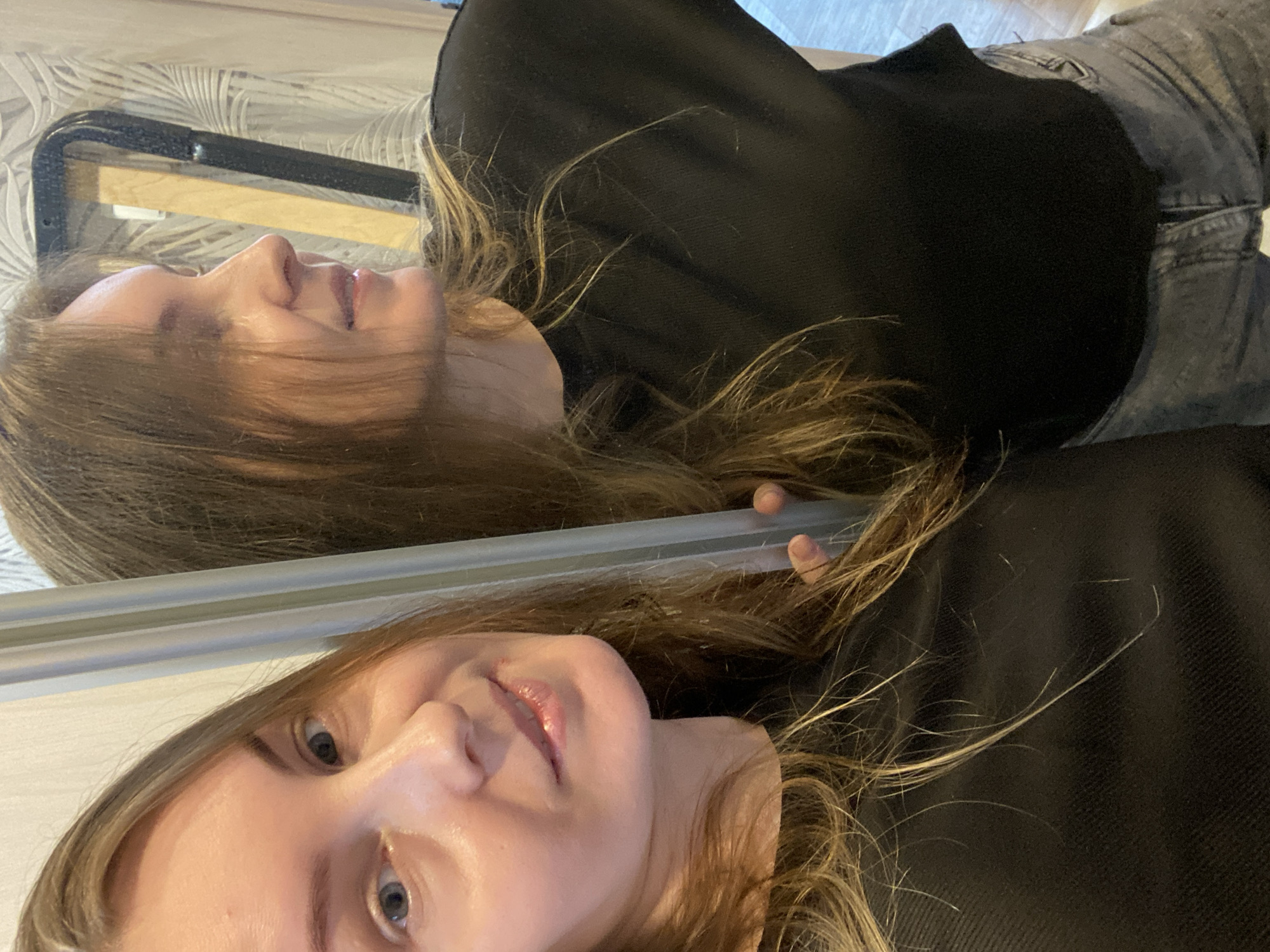A selfie-style image shows a young woman reflected in a mirror or similar reflective surface. She is wearing a black top and what appears to be gray jeans. Her hair is long and light brown with blonde highlights. The background behind her reflection includes a bed frame. No specific details indicate a location in Noviy, Russia.