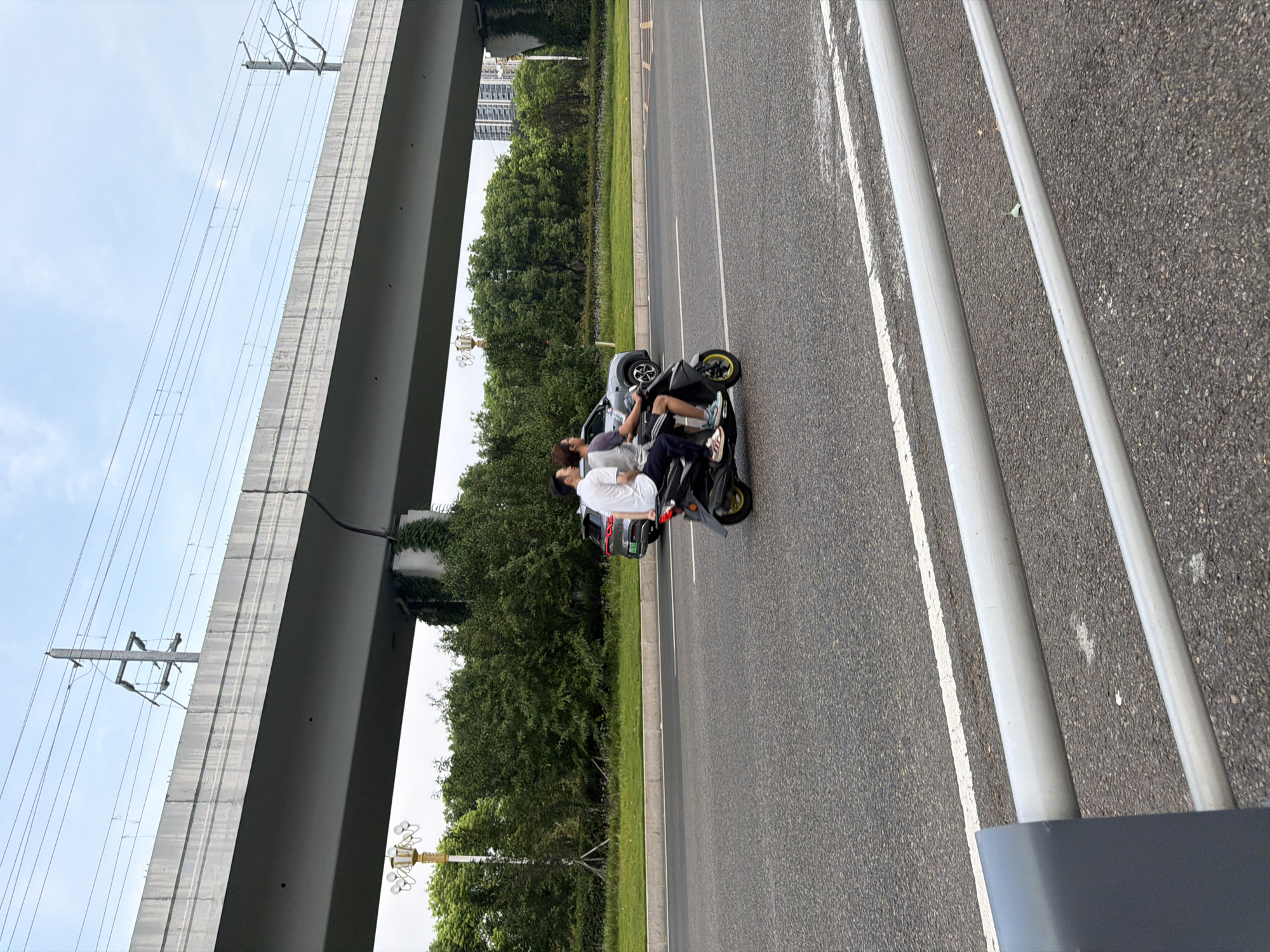 The image captures a street scene in Jiangjin, China, under a large overpass. Two individuals are riding on a dark-colored scooter. The person in the front is wearing a white polo shirt and dark shorts, while the person in the back is wearing a gray t-shirt and dark pants. They are both looking forward, and the scooter is in motion on an asphalt road. To the right of the scooter, a dark-colored SUV is parked on the side of the road. In the background, there is lush green vegetation and tall buildings, indicating an urban environment. Overhead, the concrete structure of the overpass is visible, with power lines running above it. The sky is overcast with a light blue hue. A silver metal railing is in the foreground, partially obscuring the view of the road.