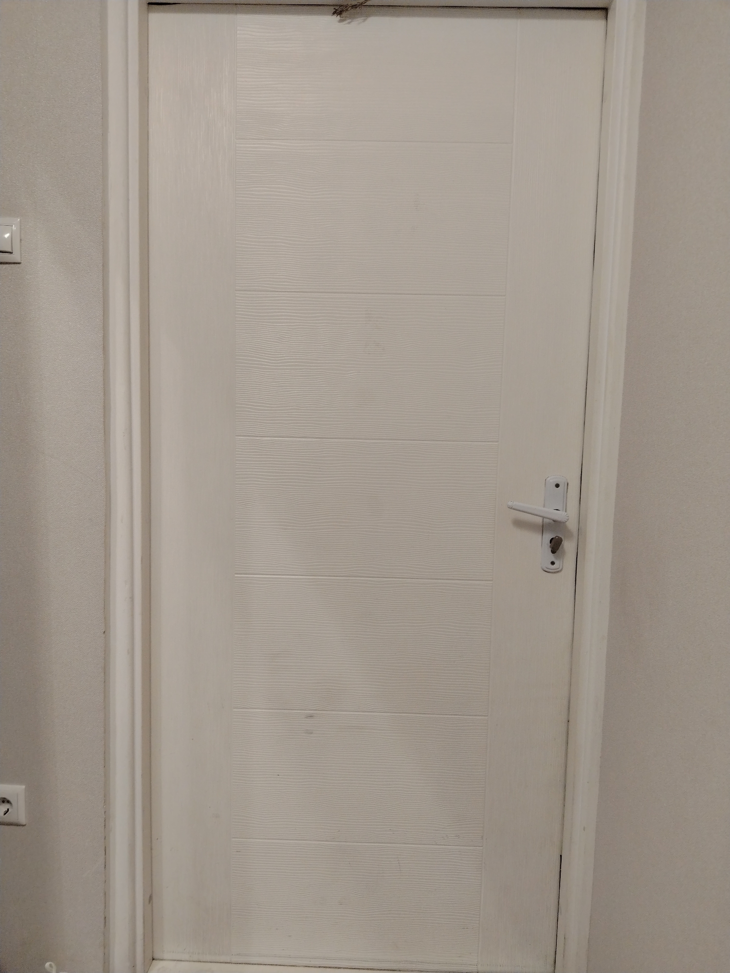 An interior full-frame vertical photograph taken in the city of Смалявічы, Belarus, displays a closed white interior door with a textured surface comprising five horizontal panels. The door is fitted with a white lever-style handle and a corresponding white keyhole lock mechanism, positioned on the right side of the door. The white door frame is visible, bordering the door on both sides. The surrounding walls are light-colored and textured. On the left wall, a white light switch with a small illuminated indicator is present in the upper-left quadrant, and a white European-style electrical outlet is partially visible in the lower-left corner. A dark smudge is evident on the upper edge of the door.