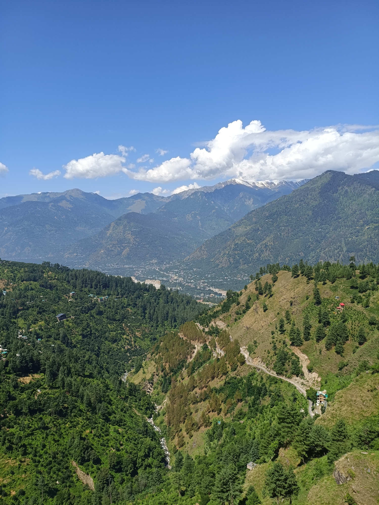 This is a high-angle, full shot of a mountainous landscape near Damadi, India, on a clear day with scattered white clouds. The image is dominated by rolling green hills covered in dense pine forests. In the foreground, a steep, grassy slope features scattered pine trees and a winding dirt road that leads to a cluster of buildings. A small red-roofed structure is visible on the hillside.
Further back, a valley opens up, revealing a small town or village nestled among the trees. The town appears to have residential buildings and some infrastructure. Beyond the valley, majestic, layered mountains rise, their peaks dusted with snow, indicating a high altitude. The sky above is a vibrant blue, with several fluffy white clouds adding texture to the scene. The overall impression is one of natural beauty and tranquility.