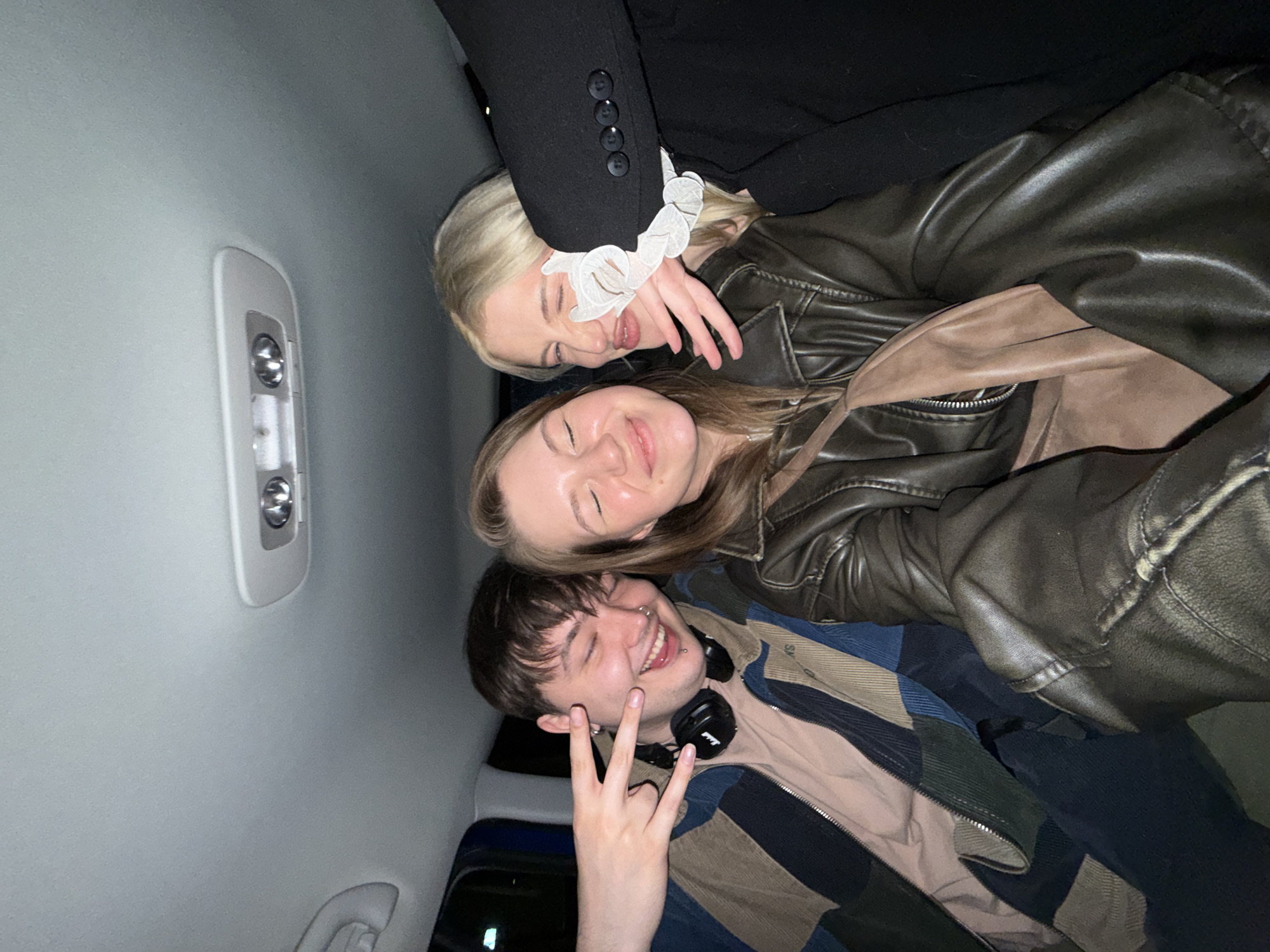 The image depicts three individuals in the interior of a vehicle, potentially a car, as suggested by the overhead lighting panel. The interior is predominantly grey. A young man with dark hair is positioned towards the bottom of the frame and is gesturing with his hand, creating a "peace" or "victory" sign. He is wearing headphones around his neck and appears to be smiling. Above him is a woman with brown hair, also smiling with her eyes closed. A blonde woman is situated at the top, with a ruffled white collar visible. Part of a black jacket with buttons is also present. The location, Belgrade, Serbia, cannot be definitively determined from the visual information present in the image.