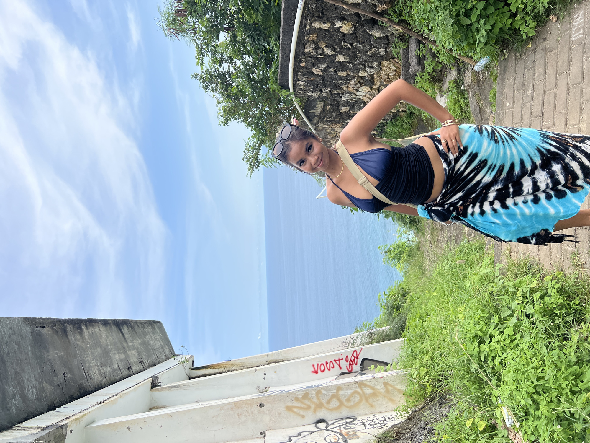 A young woman is seen in a medium, angled shot. She stands on a concrete path or walkway. The background reveals a distant expanse of ocean and a partly cloudy sky. The location is Pecatu, Indonesia. Graffiti can be seen on the nearby concrete structure, including illegible tags and the word "Victory" in red. The woman is posing, with her right hand on her hip. She wears sunglasses atop her head and is wearing a navy-blue swimsuit-style top, a crossbody strap, and a blue and black sarong. Dense green vegetation borders the path.