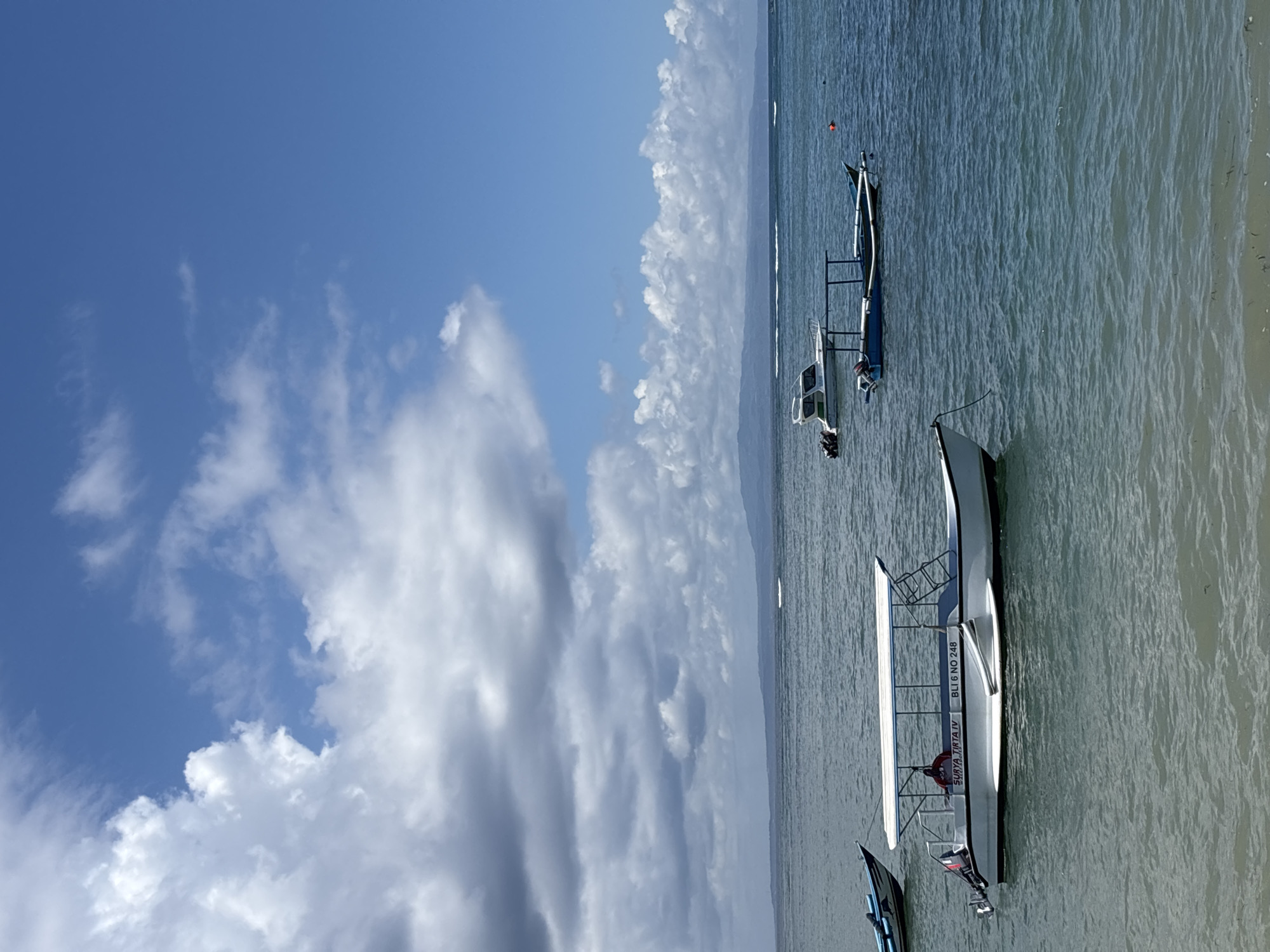 The image shows a coastal scene, likely near Denpasar, Indonesia, due to the presence of boats and the tropical setting. The sky is partially covered with white, puffy clouds against a blue background. The ocean is calm, with a light blue-green hue near the shore. Several boats are visible, some moored near the beach. One boat has the writing "SURYA TIRTA IV BLI 6 NO 248" on its side. The photo is taken at an angle, making the horizon appear vertical. There are no people visible in the image. The environment suggests a tourist location, with boats likely used for excursions or transport.