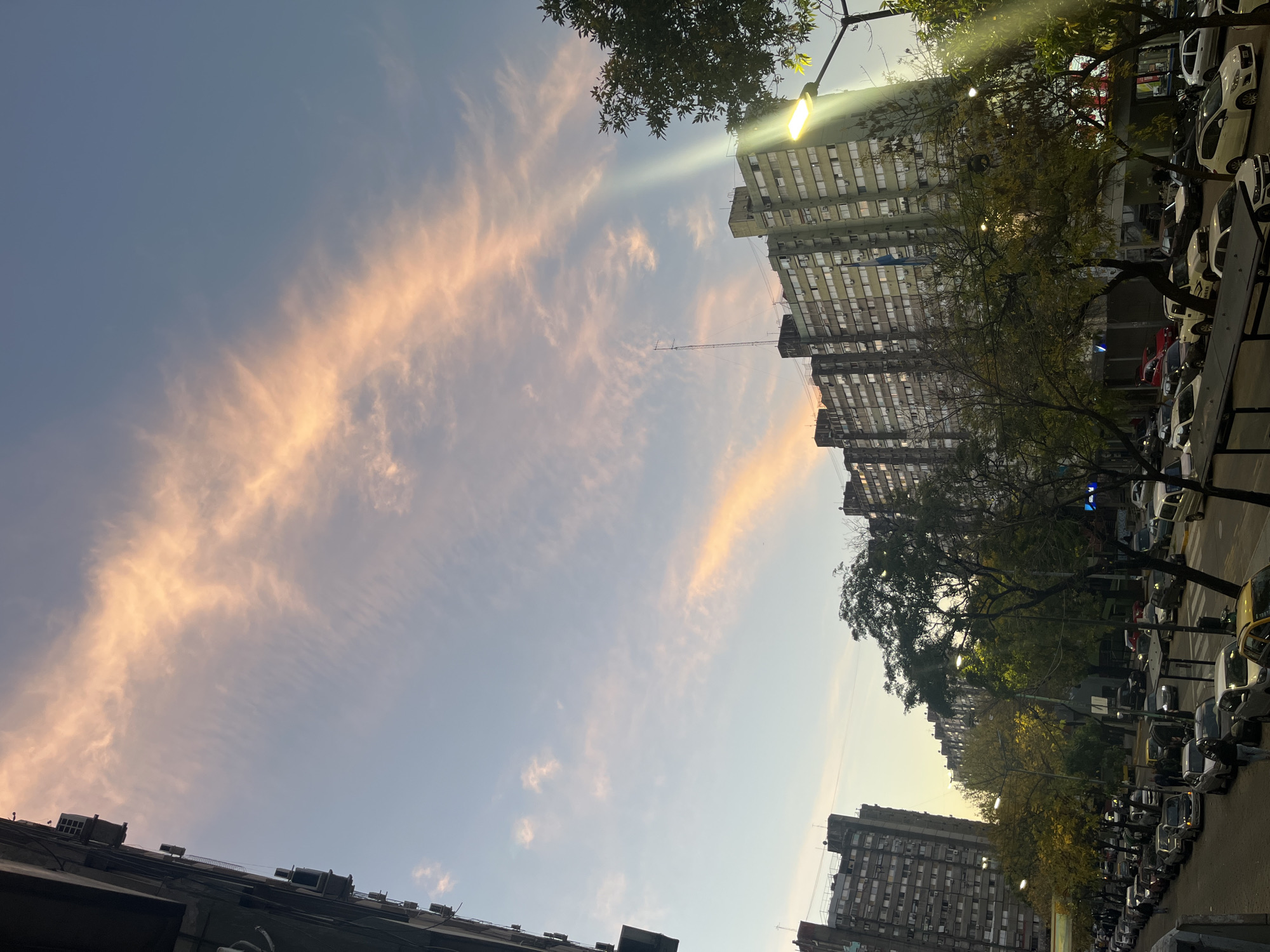 This rotated image captures an urban street scene during twilight, likely sunrise or sunset, in what is contextually identified as Buenos Aires, Argentina.
The sky occupies a significant portion of the frame, showcasing elongated, wispy clouds painted with soft pink and orange hues, contrasting against a light blue-grey background. These colors suggest the low angle of the sun.
On both sides of the street, tall, multi-story buildings with numerous windows and balconies line the view. These structures appear to be made of concrete or similar light-colored materials, some showing signs of weathering. From one of the buildings, a slender antenna or mast extends towards the center of the sky.
Green trees with varying densities of foliage are visible, lining the street and interspersed among the buildings. Bright streetlights are active; one particularly luminous lamp on the right side casts a strong, upward beam of white light, illuminating the upper parts of the buildings and emphasizing the evening or early morning ambiance.
The street below the buildings is filled with numerous parked cars, many of which are light-colored or white. Closer inspection reveals that some of these vehicles display the word "UBER" on their rooftops, indicating ride-sharing services or taxis are present. No other significant text is legible on signs or other objects within the image.
The overall scene presents a blend of natural beauty in the sky and the bustling, illuminated infrastructure of a city at a transitional time of day.